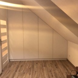 Stanislav Alimgulov - Biała zabudowa meblowa pod skosem na poddaszu, z widocznymi drzwiami po lewej stronie i drewnianą podłogą.