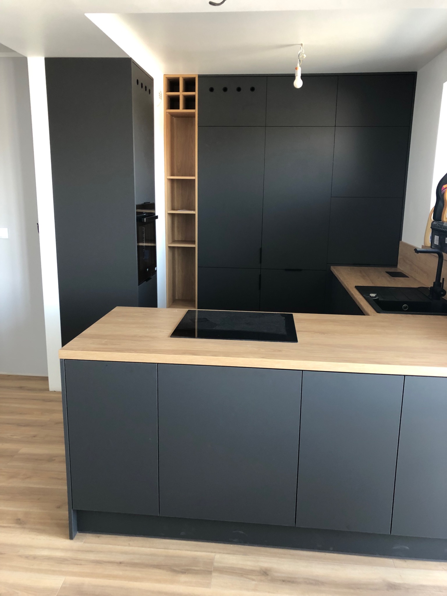 Nowoczesna kuchnia z frontami w kolorze grafitowym, drewnianym blatem i otwartymi półkami z jasnego drewna, oświetlenie LED w pionowej wnęce.