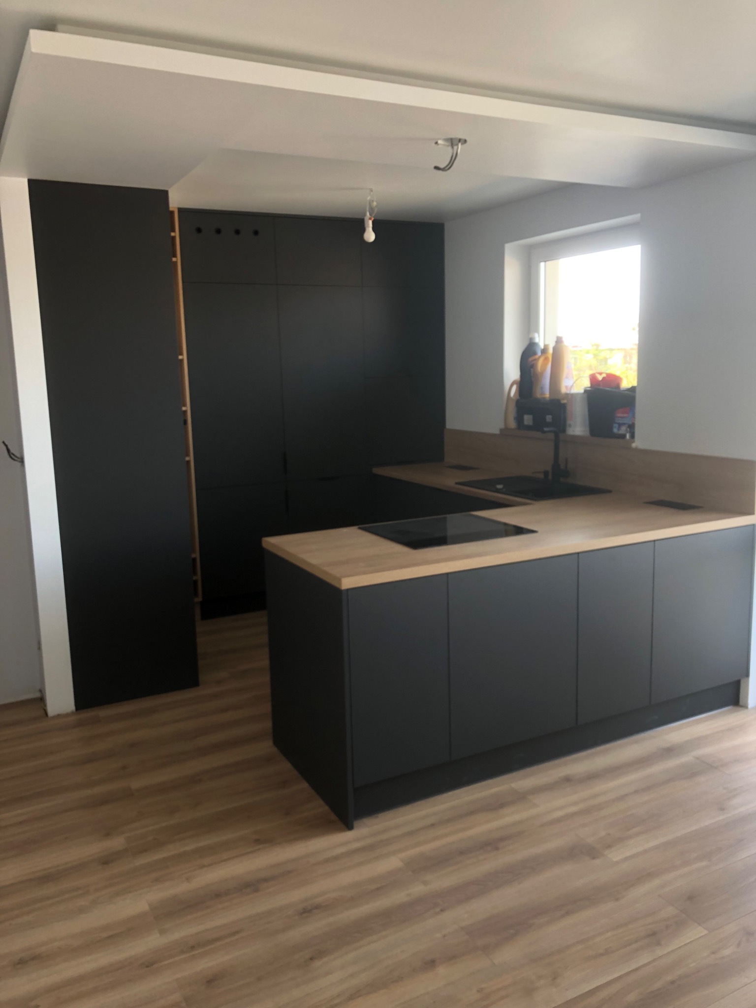 Nowoczesna kuchnia w odcieniach czerni i drewna, z minimalistycznymi frontami szafek i drewnianym blatem, oświetlona naturalnym światłem z okna, z widocznymi akcesoriami na blacie i drewnianą podłogą.