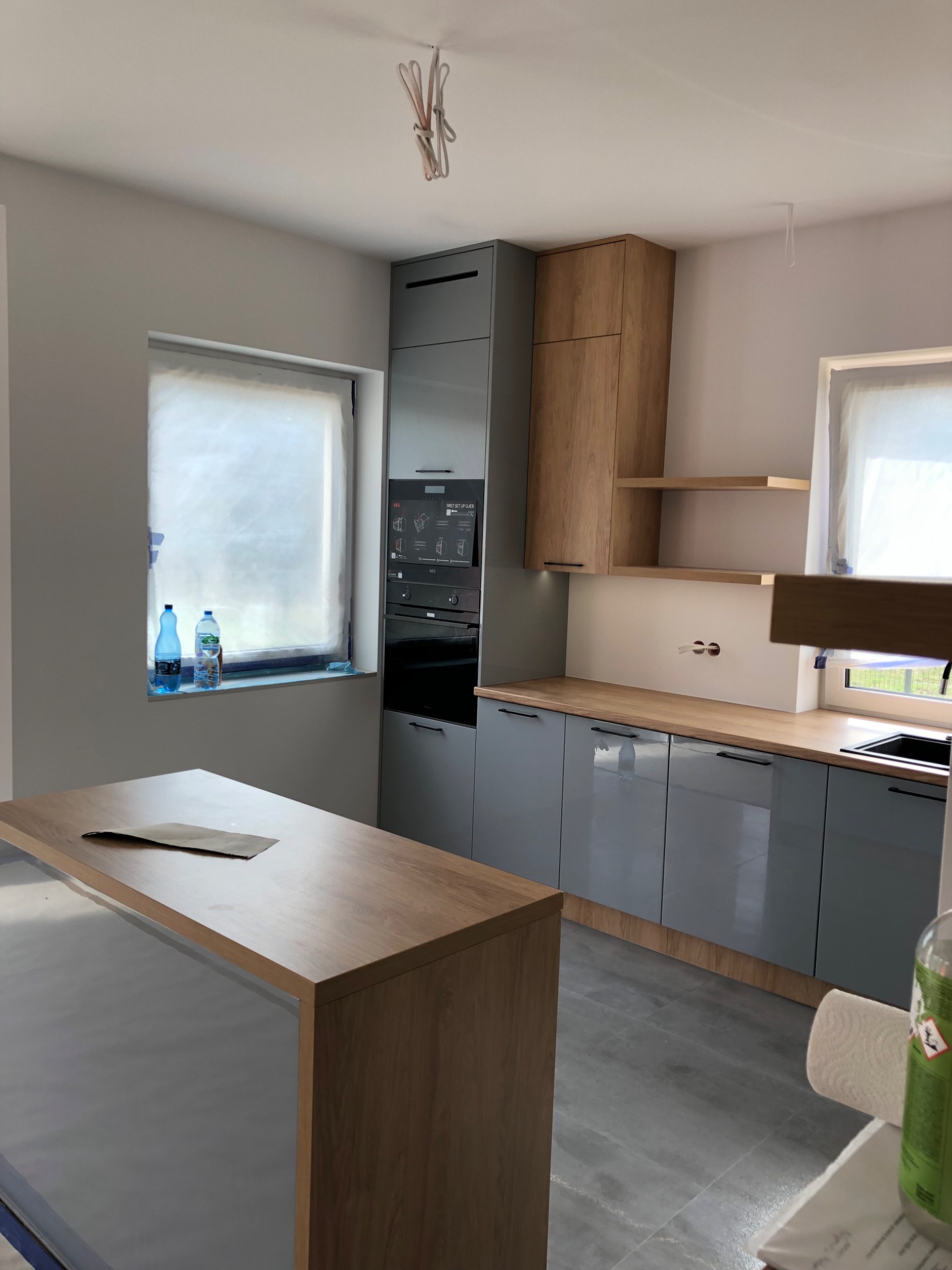 Szara kuchnia na wymiar z drewnianymi akcentami, wysoka zabudowa z piekarnikiem, okno zasłonięte folią ochronną, butelki wody na parapecie, wyspa kuchenna z blatem drewnopodobnym.