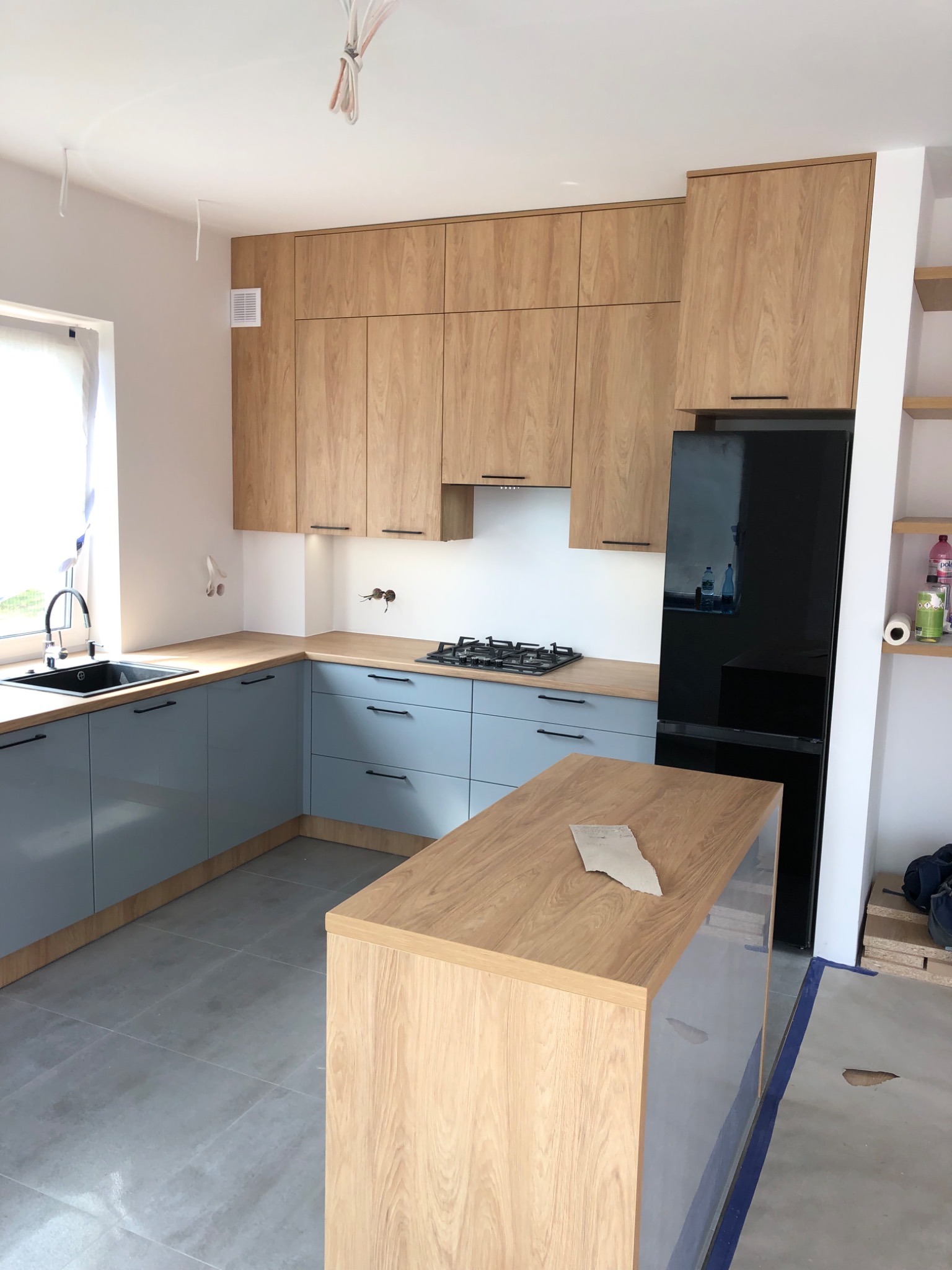 Nowoczesna kuchnia z szarymi szafkami dolnymi i drewnianymi górnymi, czarnym sprzętem AGD, widoczne niedokończone prace wykończeniowe.