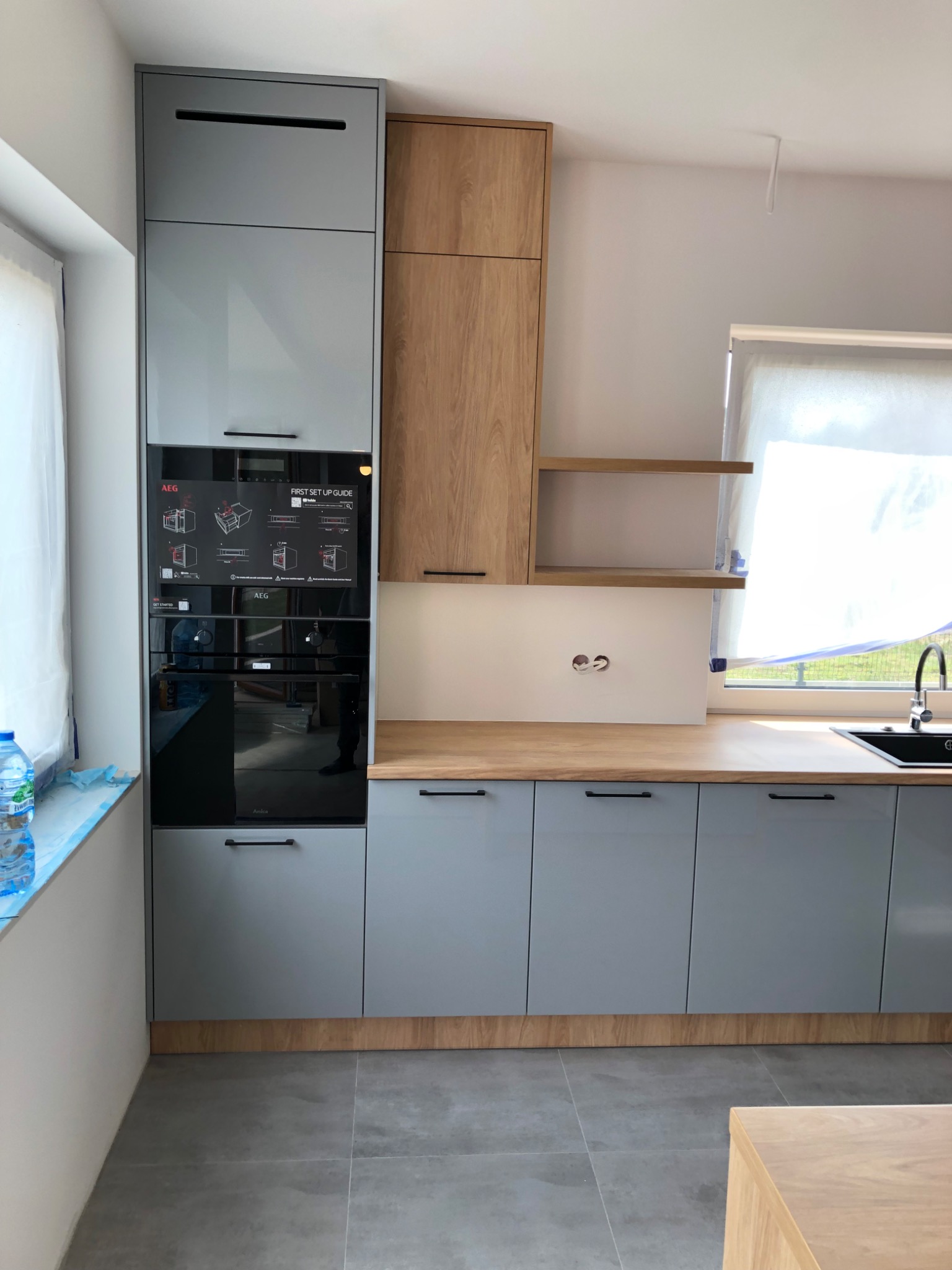Nowoczesna zabudowa kuchenna z szarymi lakierowanymi frontami, drewnianymi akcentami i wbudowanym piekarnikiem AEG, widoczne instrukcje obsługi na piekarniku.