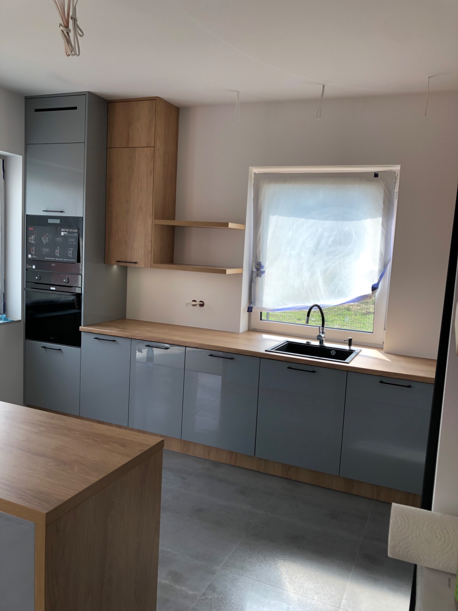 Szara zabudowa kuchenna z drewnianym blatem, widoczne sprzęty AGD w zabudowie oraz wyspa kuchenna z drewnianym blatem. Okno zaklejone folią ochronną.