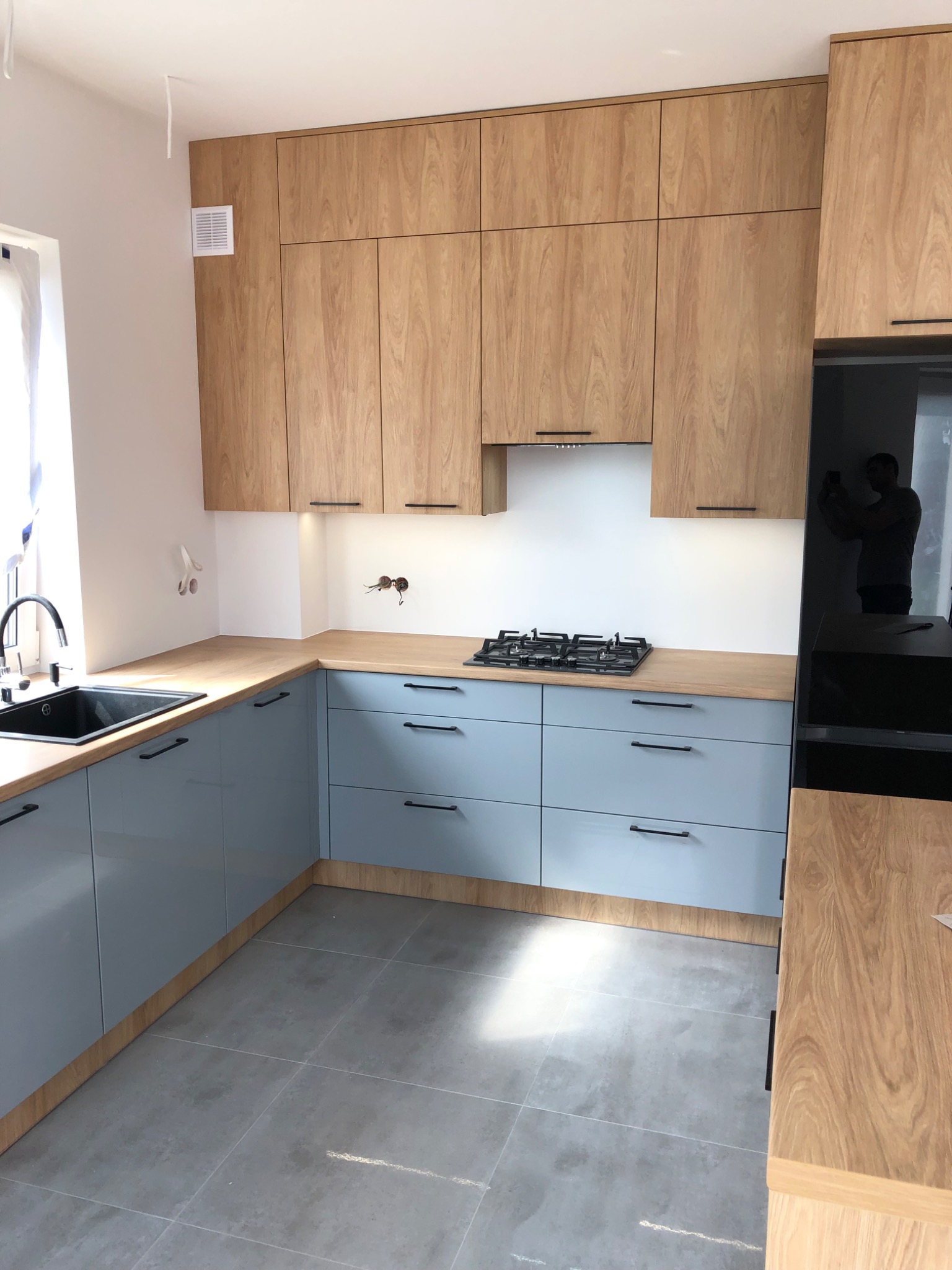 Jasna kuchnia z drewnianymi szafkami górnymi i błękitnymi dolnymi, czarny zlew i płyta gazowa, widoczne niedokończone gniazdka elektryczne.