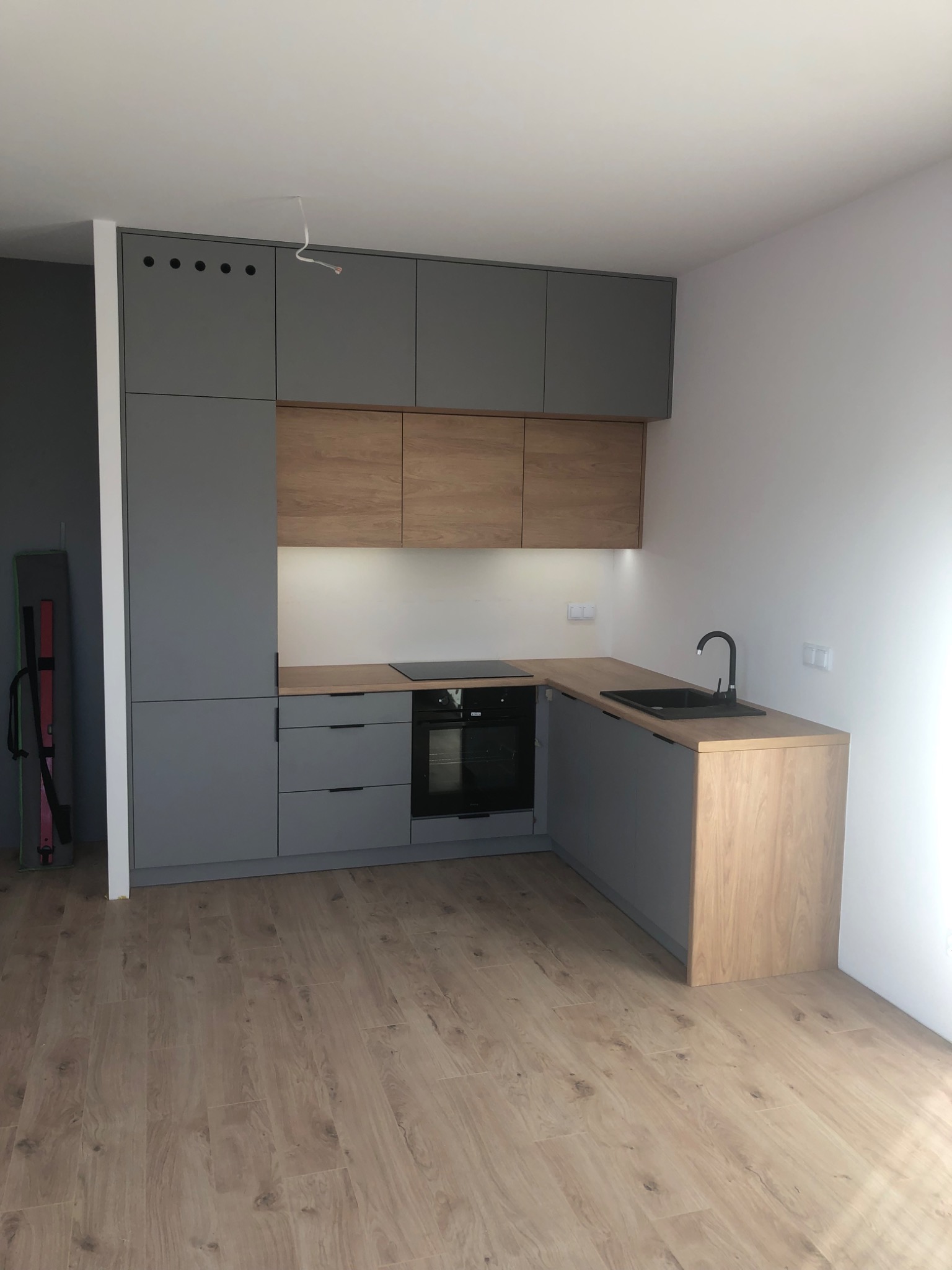 Nowoczesna kuchnia z szarymi szafkami i drewnianymi blatami, czarną armaturą i płytą indukcyjną, widok na jasną podłogę w nowym mieszkaniu.