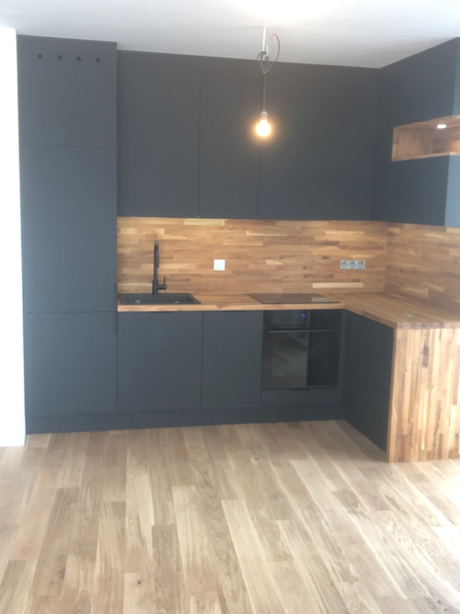 Nowoczesna kuchnia z grafitowymi szafkami i drewnianym blatem oraz panelem ściennym, oświetlona wiszącą żarówką na tle parkietu.
