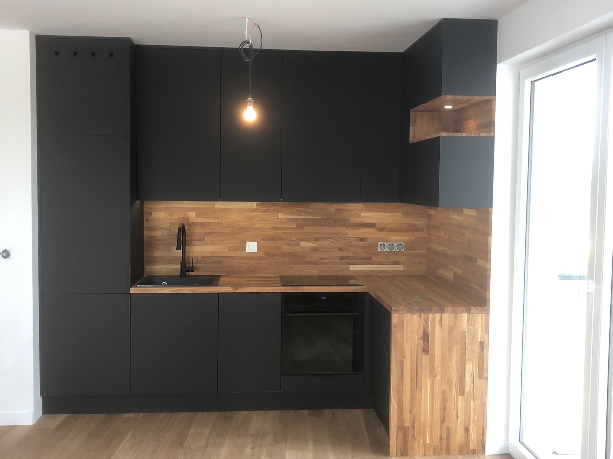 Nowoczesna kuchnia z czarnymi matowymi szafkami, drewnianym blatem i backsplashiem z desek, oświetlona wiszącą żarówką na kablu, z widoczną zabudową piekarnika i zlewozmywakiem.
