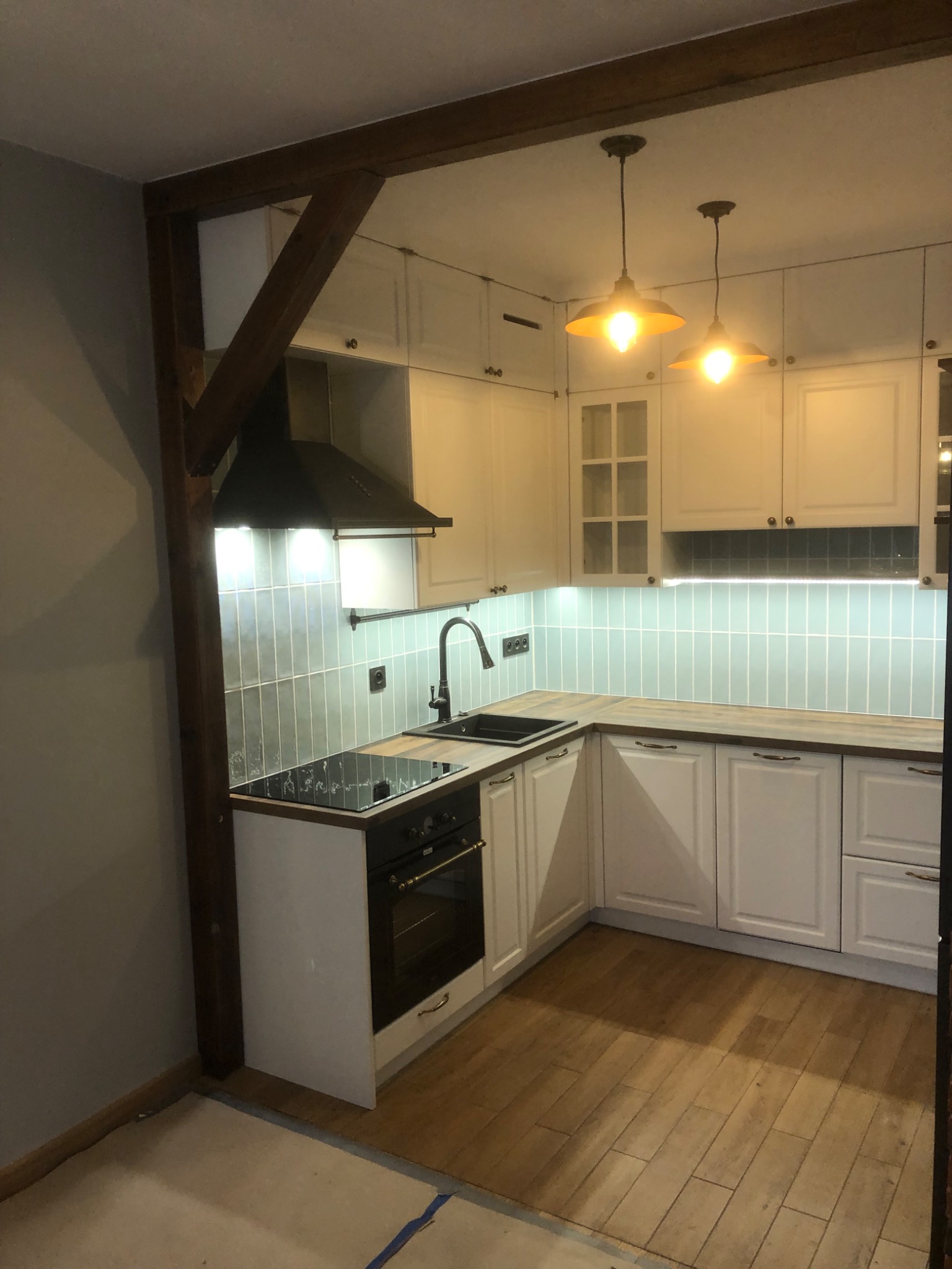 Jasna kuchnia z drewnianą konstrukcją, białymi szafkami i drewnianym blatem, oświetlona dwoma lampami w stylu retro.