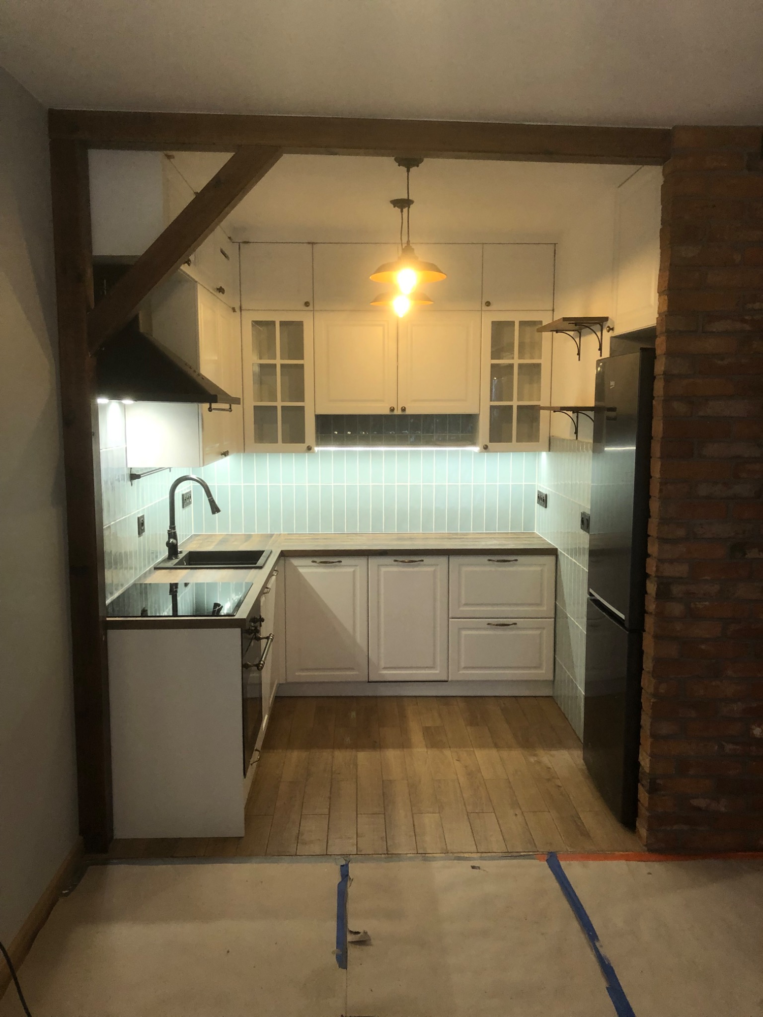 Kuchnia w stylu rustykalnym z białymi szafkami, drewnianym blatem i ceglanym filarem, oświetlona lampą wiszącą z żarówką edisona, widok z niedokończonego pomieszczenia z podłogą zabezpieczoną taśmą...