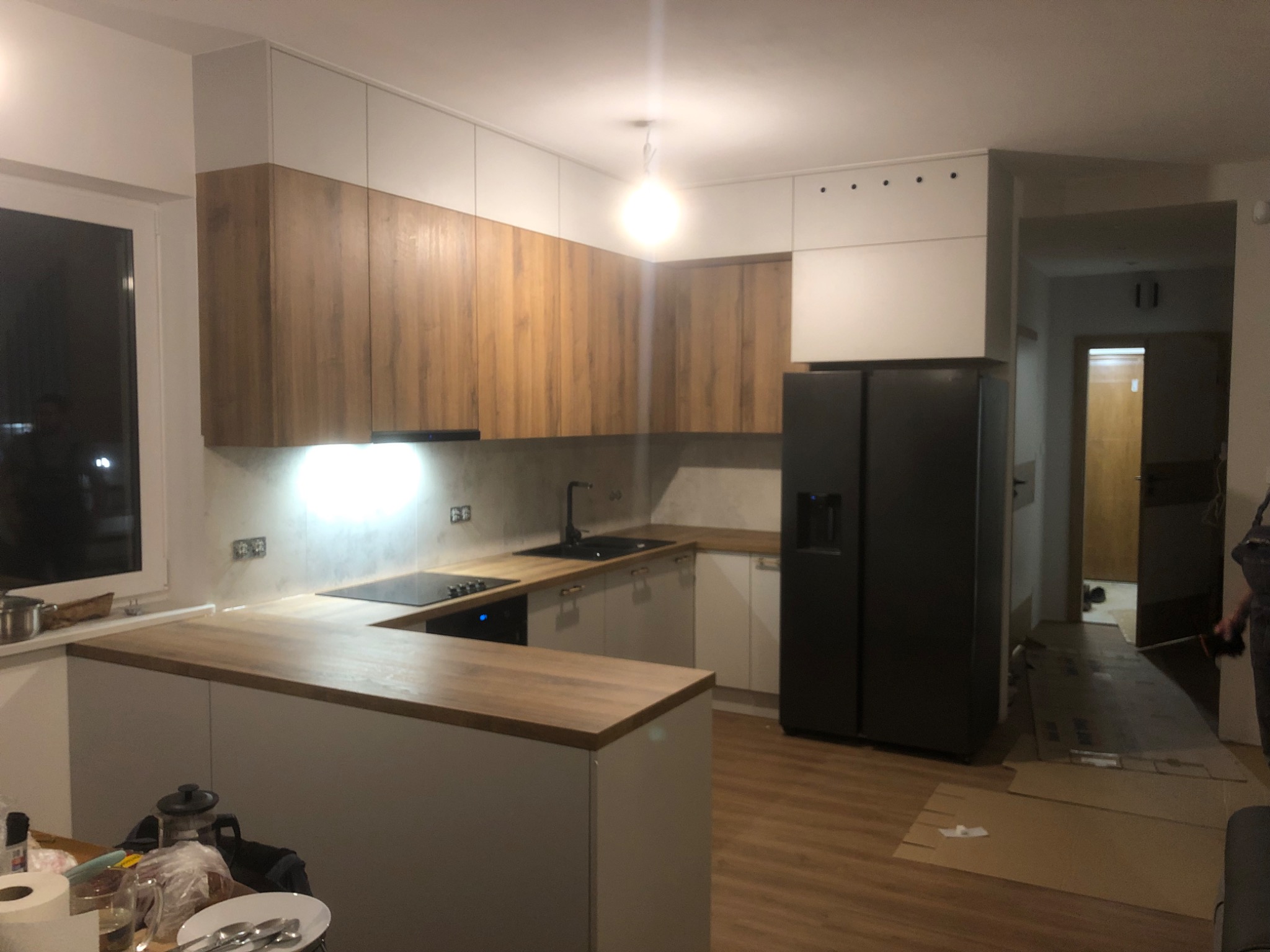 Nowoczesna kuchnia z drewnianymi frontami szafek górnych i białymi dolnymi, czarnym zlewem i płytą indukcyjną, oświetleniem podszafkowym, czarną lodówką side-by-side, częściowo rozpakowana...