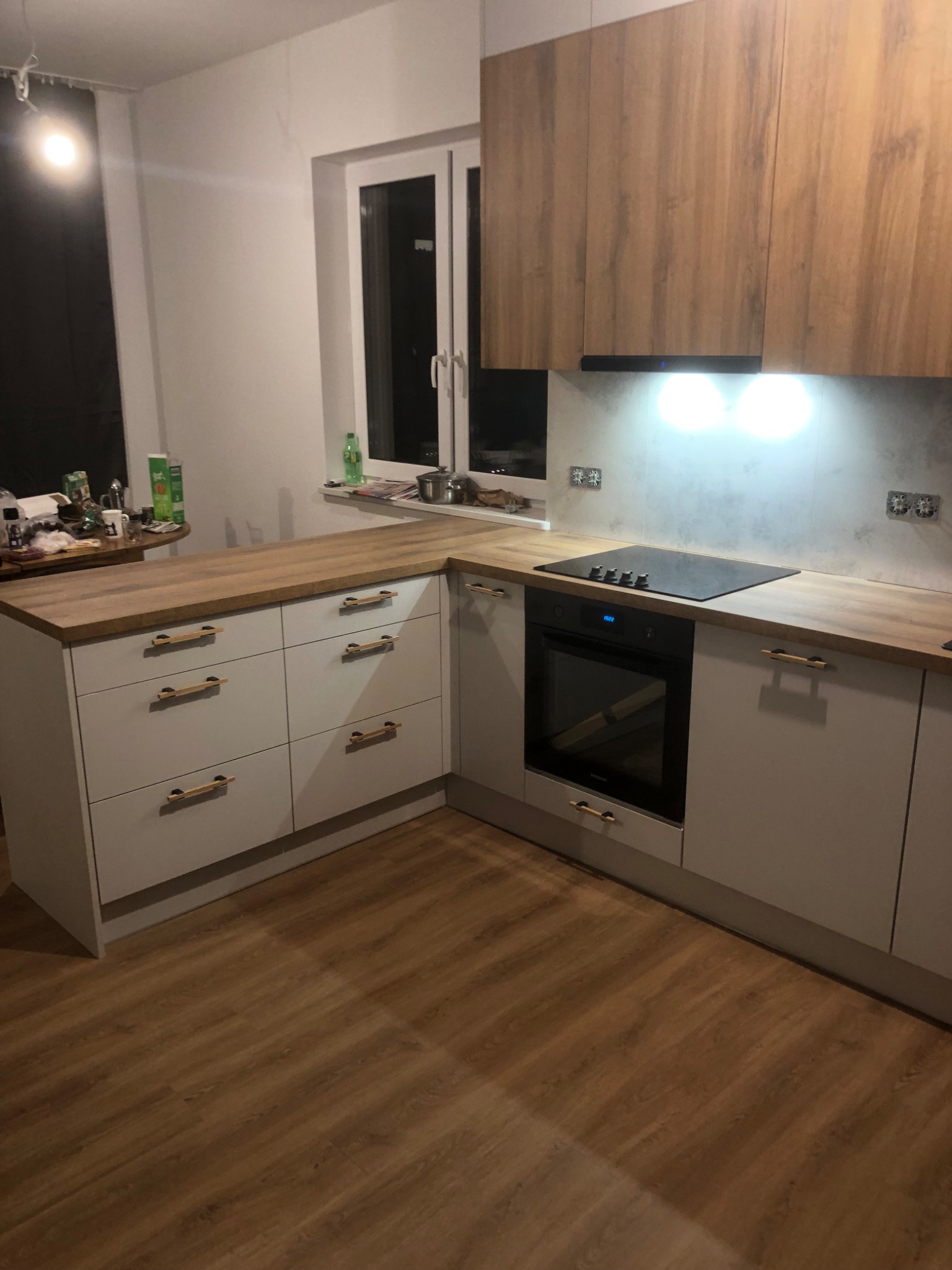 Nowoczesna kuchnia z szarymi szafkami, drewnianym blatem i złotymi uchwytami, płyta indukcyjna i piekarnik w zabudowie, podświetlenie LED nad blatem roboczym, podłoga laminowana imitująca drewno.