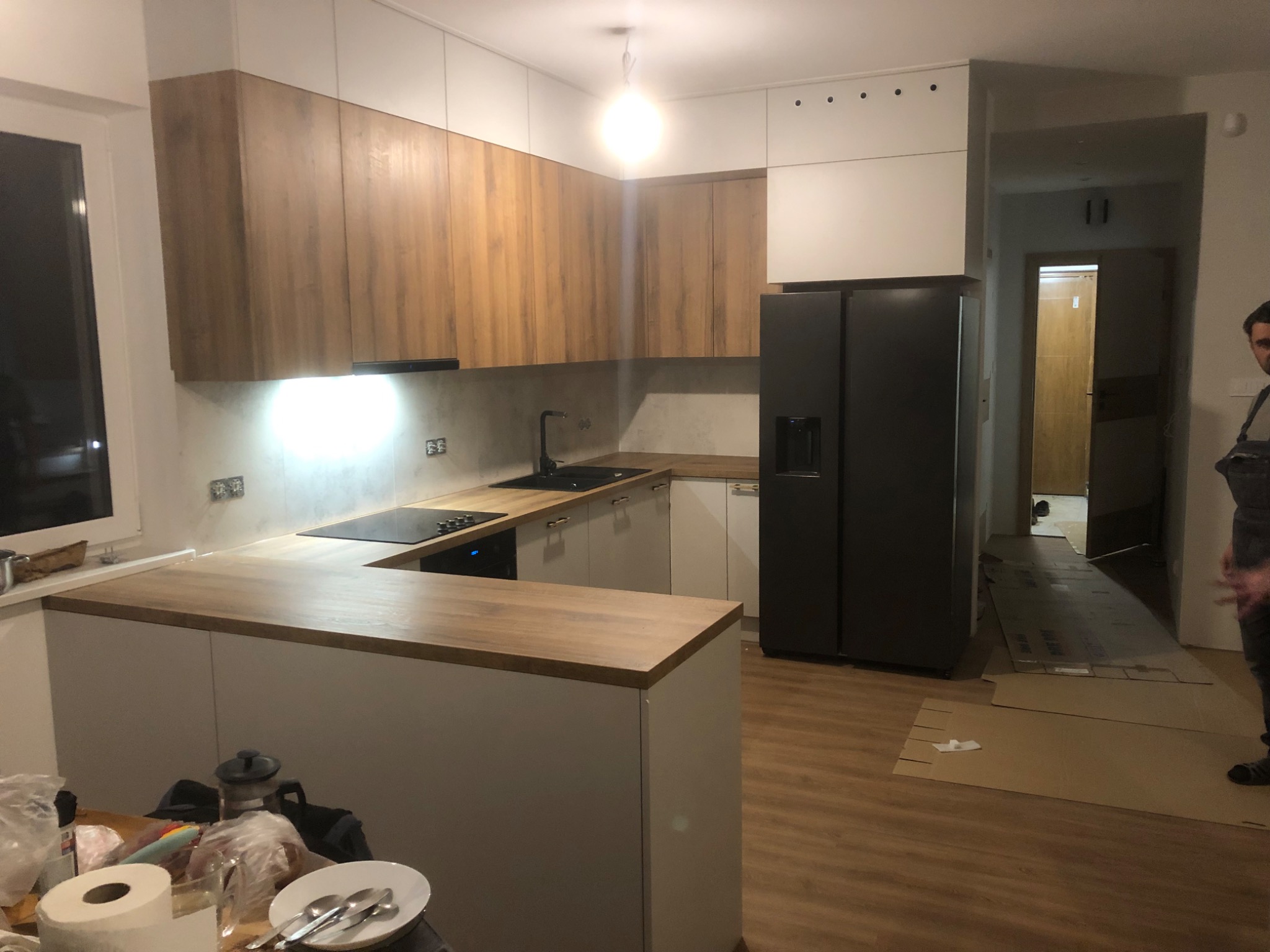 Nowoczesna kuchnia z drewnianymi górnymi szafkami i białymi dolnymi, czarnym sprzętem AGD i blatem z drewnopodobnego laminatu, widoczny fragment osoby w roboczym ubraniu oraz resztki po posiłku...