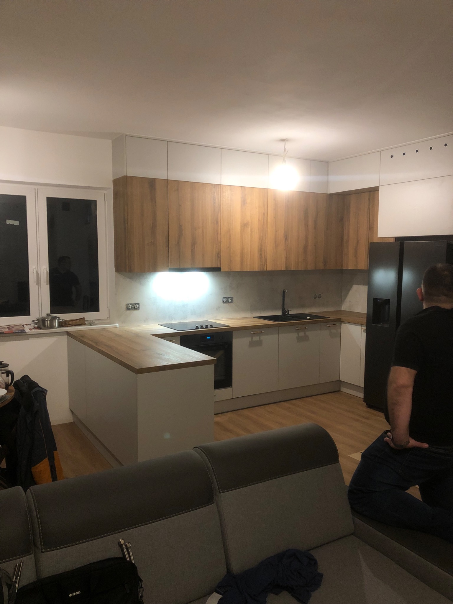 Nowoczesna kuchnia z drewnianymi szafkami górnymi i białymi dolnymi, wyspa z drewnianym blatem, czarna lodówka side-by-side, widok z salonu na częściowo umeblowaną przestrzeń.