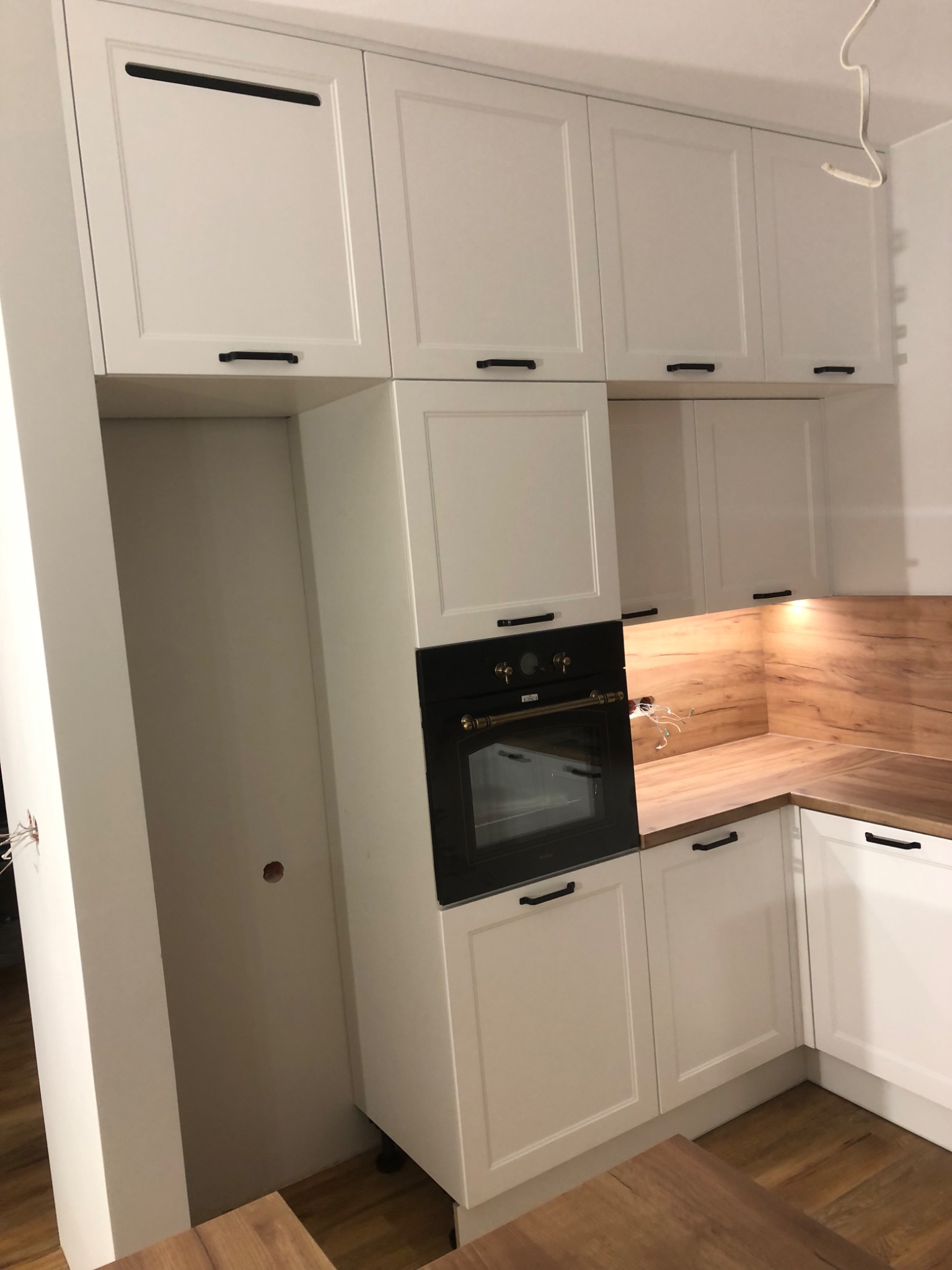 Biała zabudowa kuchenna z czarnym piekarnikiem, drewnianym blatem i podświetleniem, widoczne kable wystające ze ściany, wnęka na lodówkę.