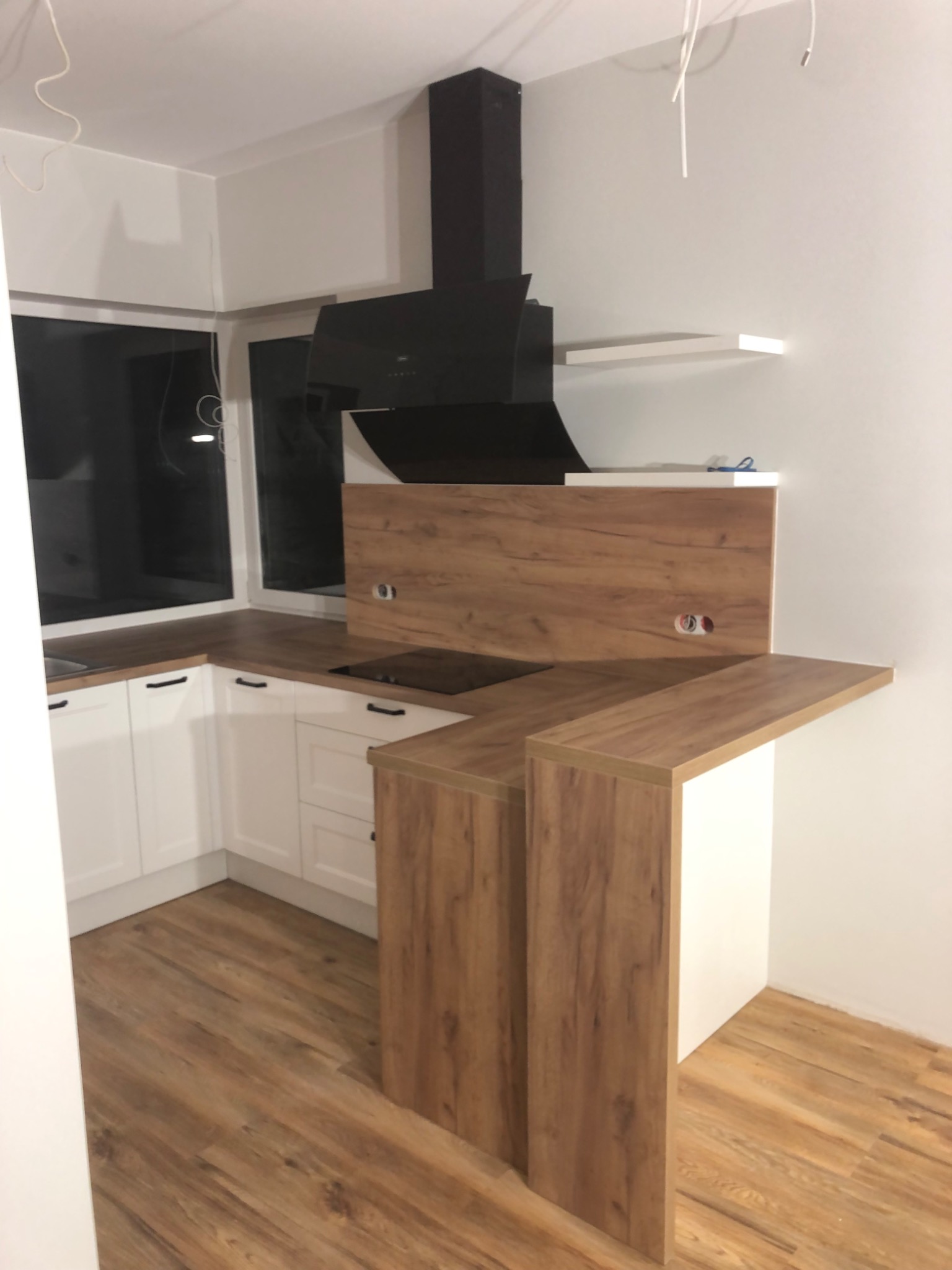 Nowoczesna kuchnia z białymi szafkami i drewnianymi blatami, czarny okap, drewniana podłoga, widoczne gniazdka elektryczne w panelu nad blatem.