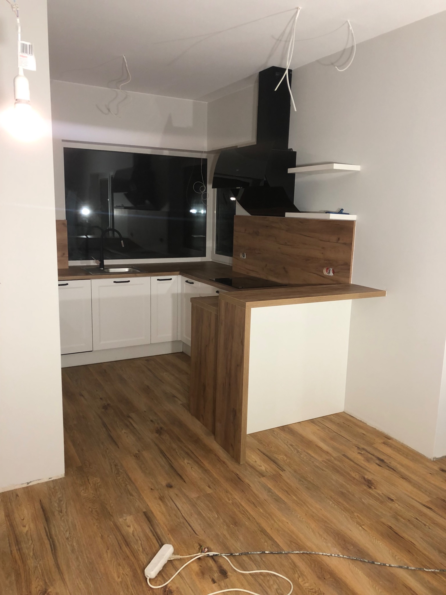 Wnętrze kuchni w trakcie montażu: białe szafki, drewnopodobny blat, czarny okap, niedokończone podłączenia elektryczne zwisające z sufitu, widoczne gniazdka na ścianie.