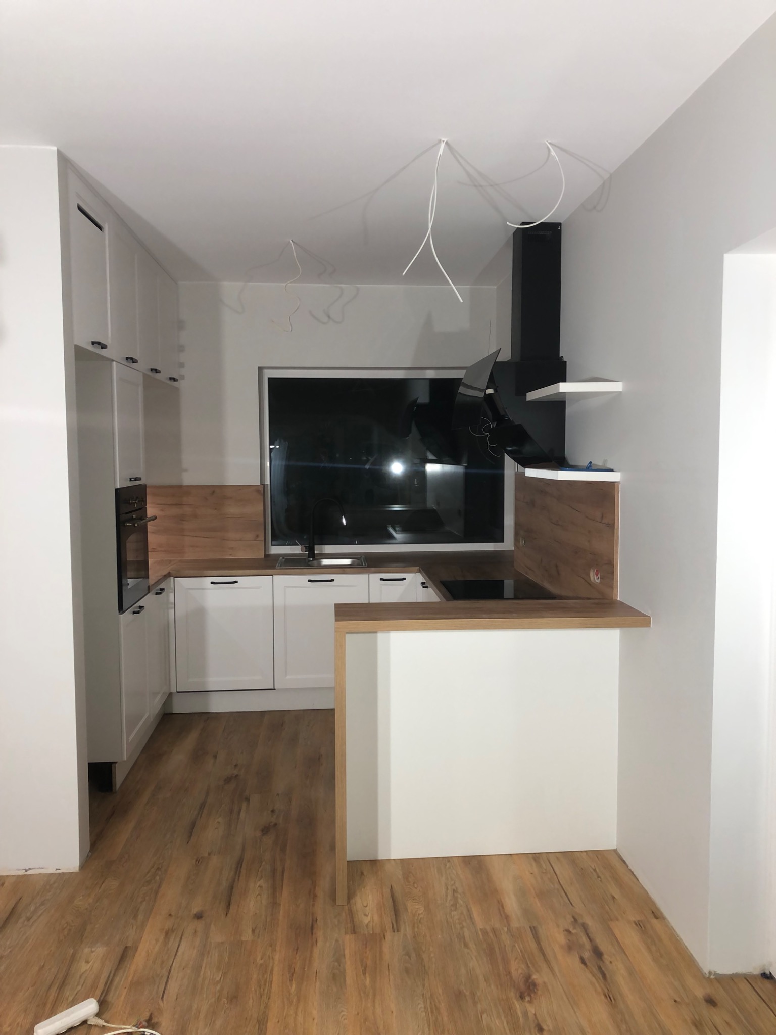 Nowoczesna kuchnia w trakcie montażu: białe szafki z czarnymi uchwytami, drewniane blaty, czarny okap, widoczne kable wystające z sufitu.