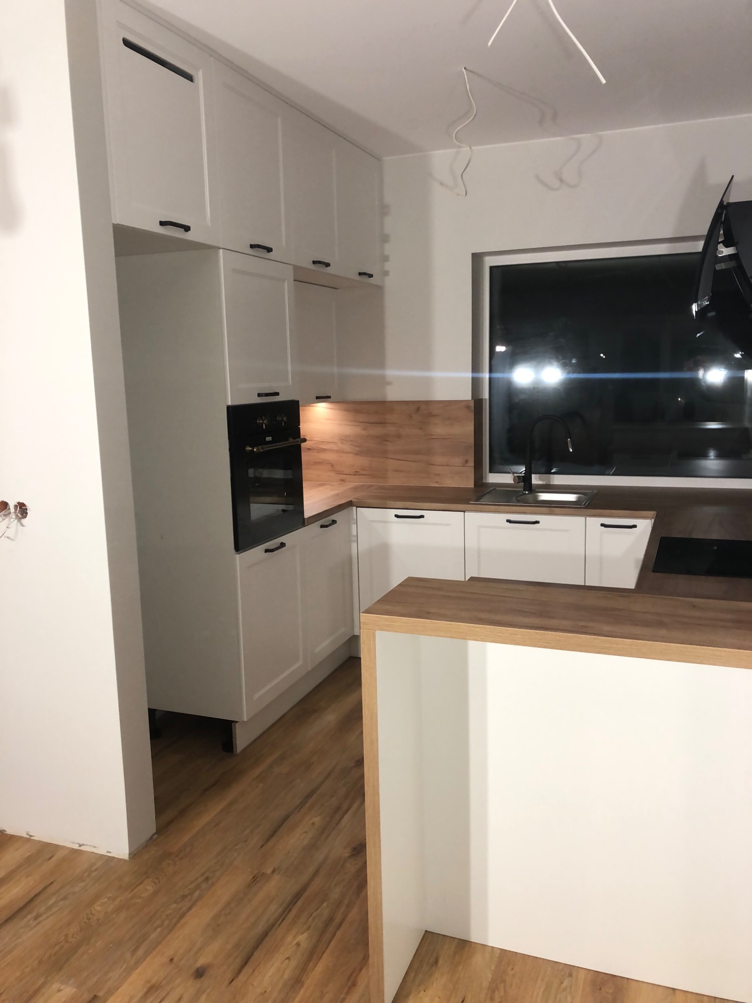 Nowoczesna, biała zabudowa kuchenna z drewnianymi blatami i czarnymi uchwytami, widoczna w trakcie montażu, z wiszącymi kablami i niedokończonymi ścianami.