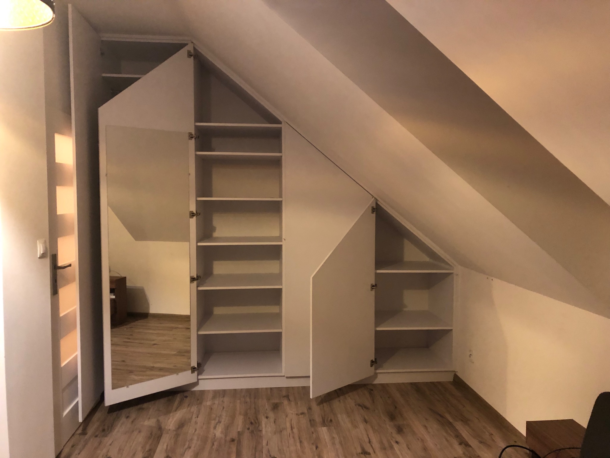 Biała zabudowa meblowa na poddaszu ze skośnym sufitem, z otwartymi półkami i drzwiami z lustrem.