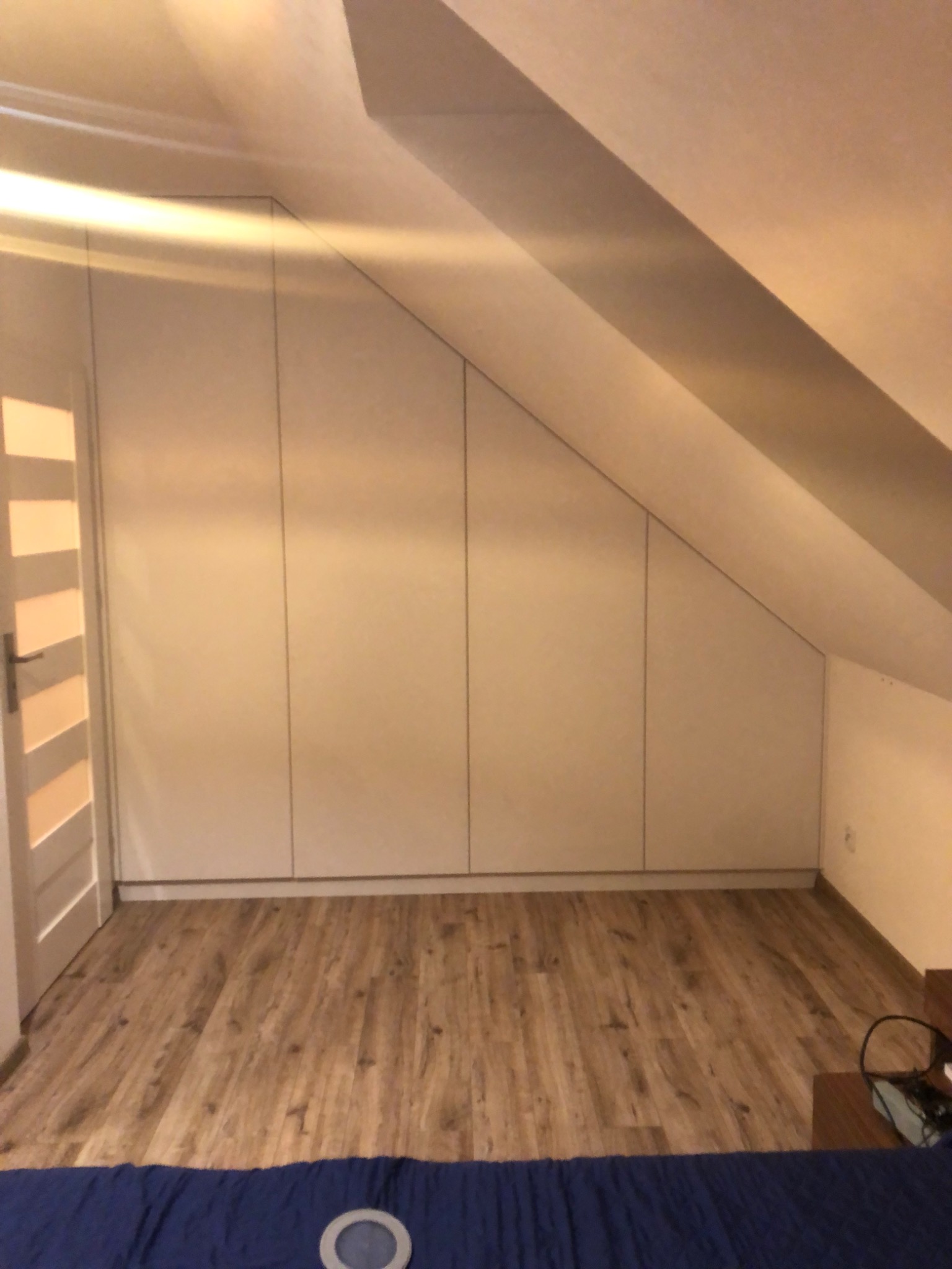 Biała zabudowa meblowa pod skosem na poddaszu, z widocznymi drzwiami po lewej stronie i drewnianą podłogą.