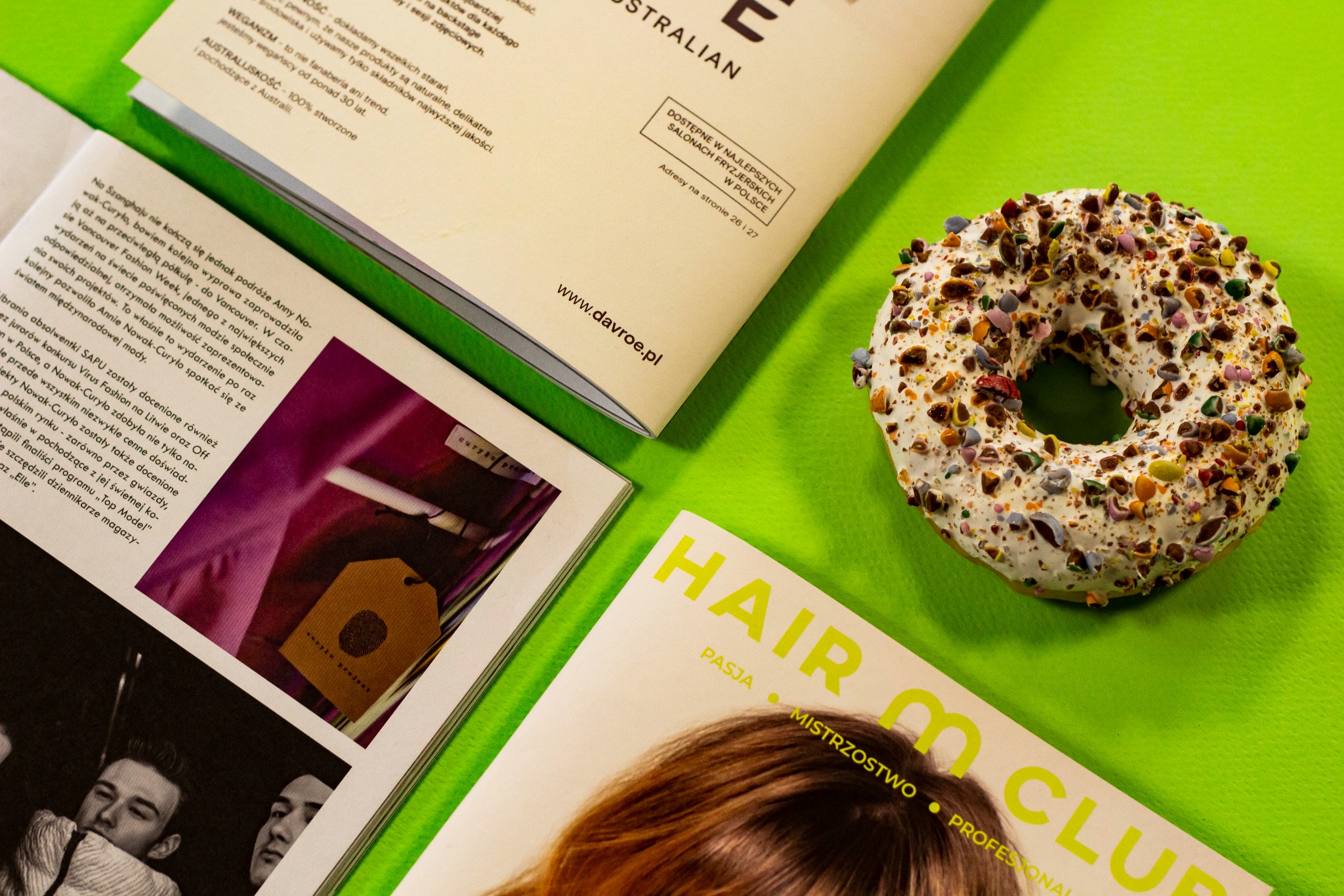 Donut polany białym lukrem i posypany kolorowymi kawałkami, leżący na zielonym tle obok otwartych magazynów z reklamami salonów fryzjerskich i artykułami o modzie.