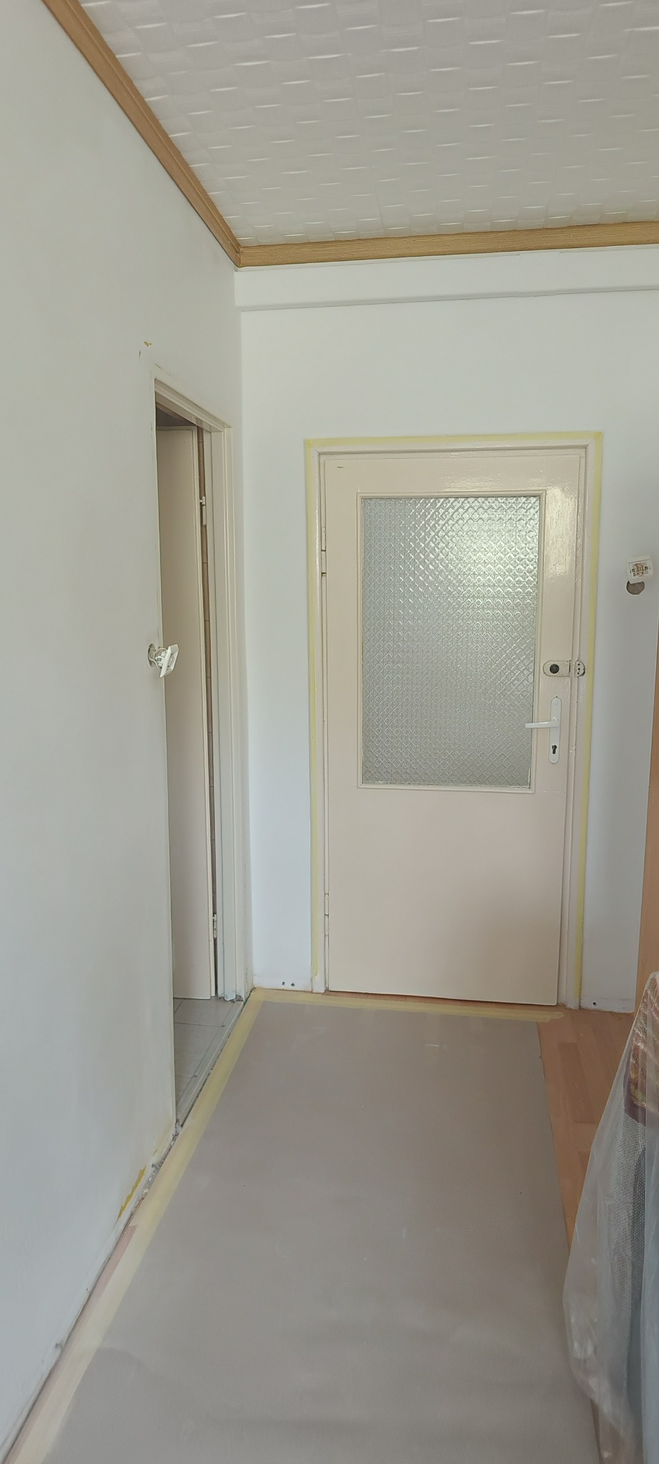 Korytarz w trakcie prac remontowych, podłoga zabezpieczona folią malarską, ściany świeżo pomalowane na biało, widoczne taśmy malarskie wokół drzwi i listew przypodłogowych.