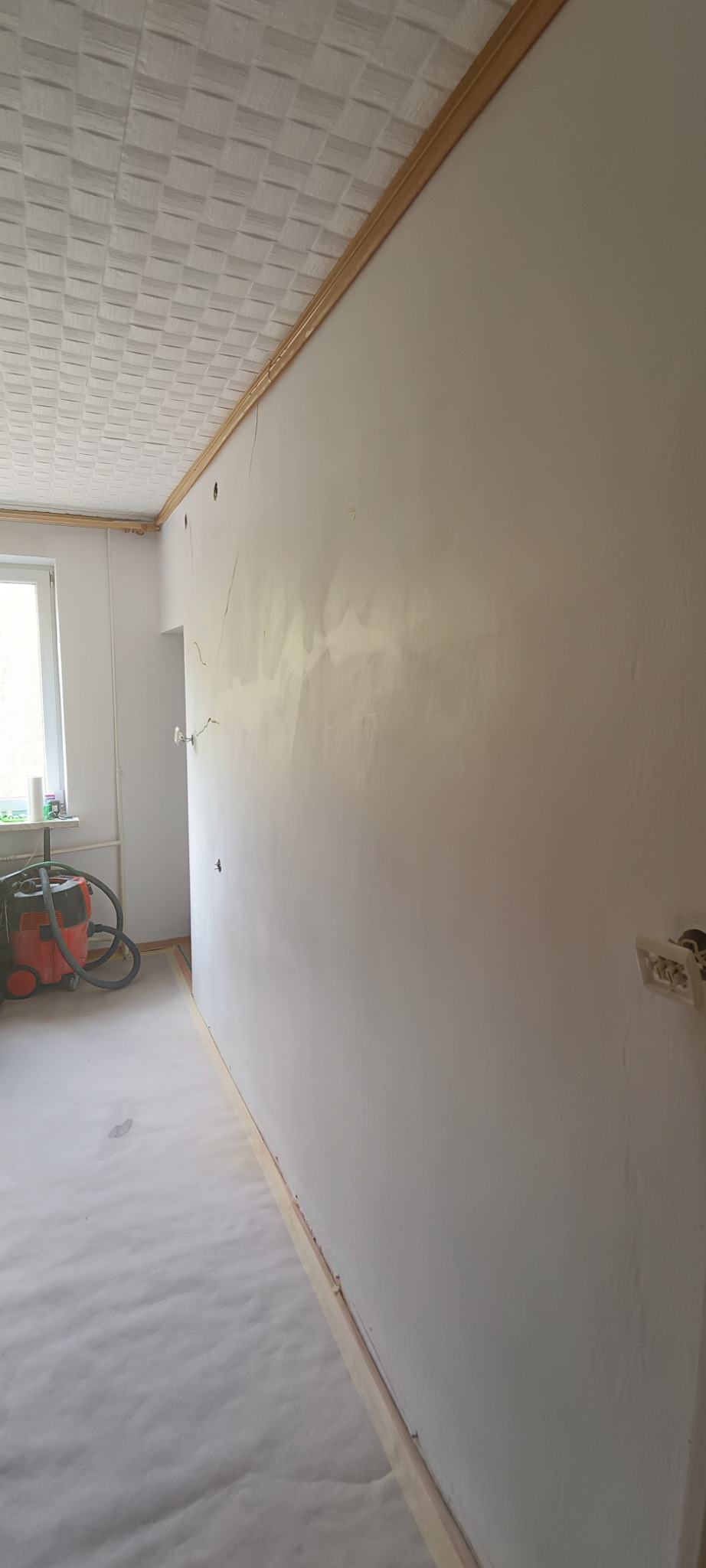 Pokój w trakcie remontu, ściana po nałożeniu gładzi, widoczne nierówności przed szlifowaniem, podłoga zabezpieczona folią, odkurzacz przemysłowy w tle.