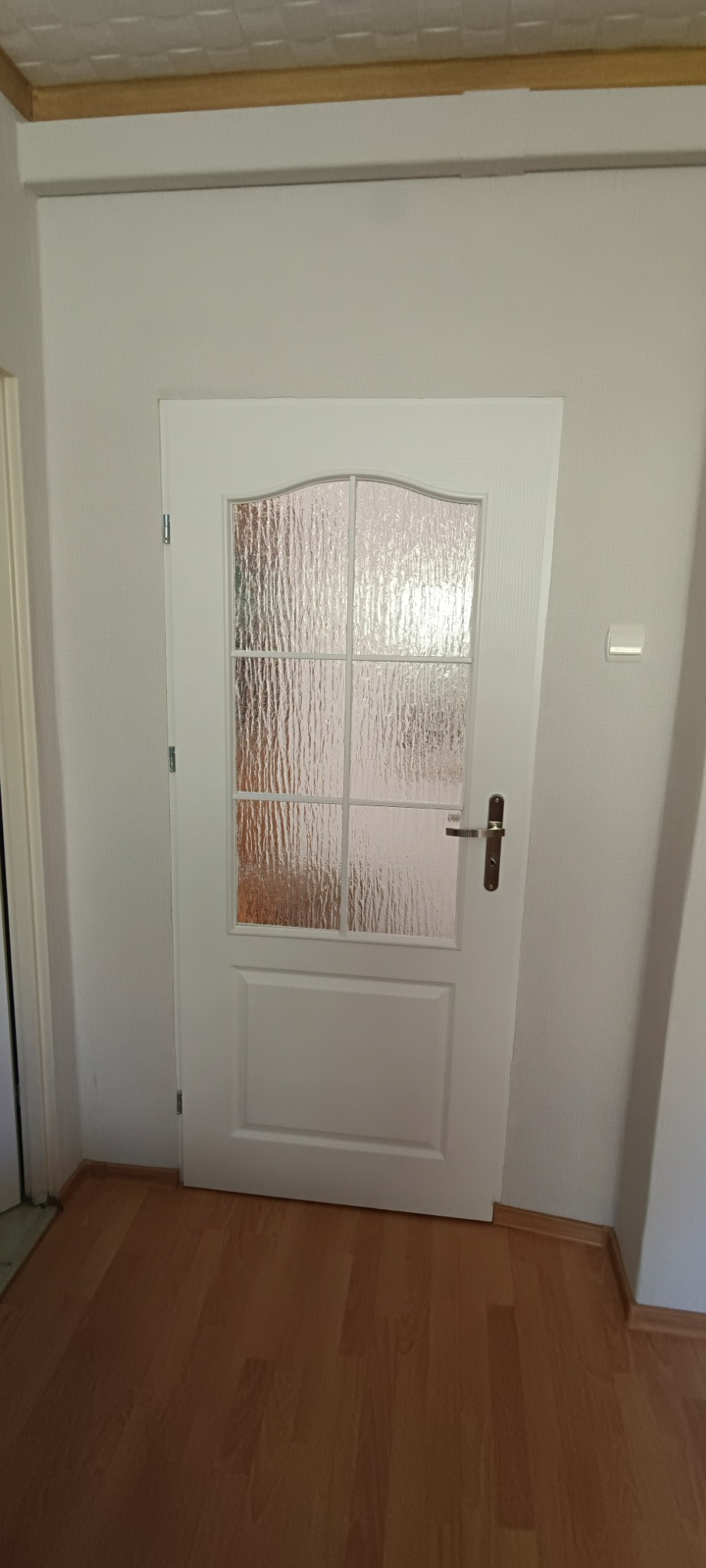 Białe drzwi wewnętrzne z szybą o fakturze lodu, zamontowane w szarej ścianie, widoczne drewniane panele podłogowe.