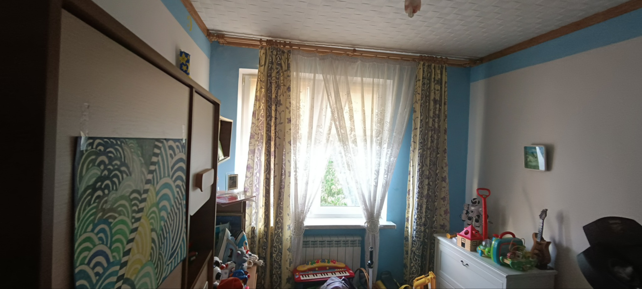 Pokój dziecięcy z jasnymi ścianami, zasłoniętym oknem, grzejnikiem pod oknem i zabawkami na komodzie oraz przy ścianie.