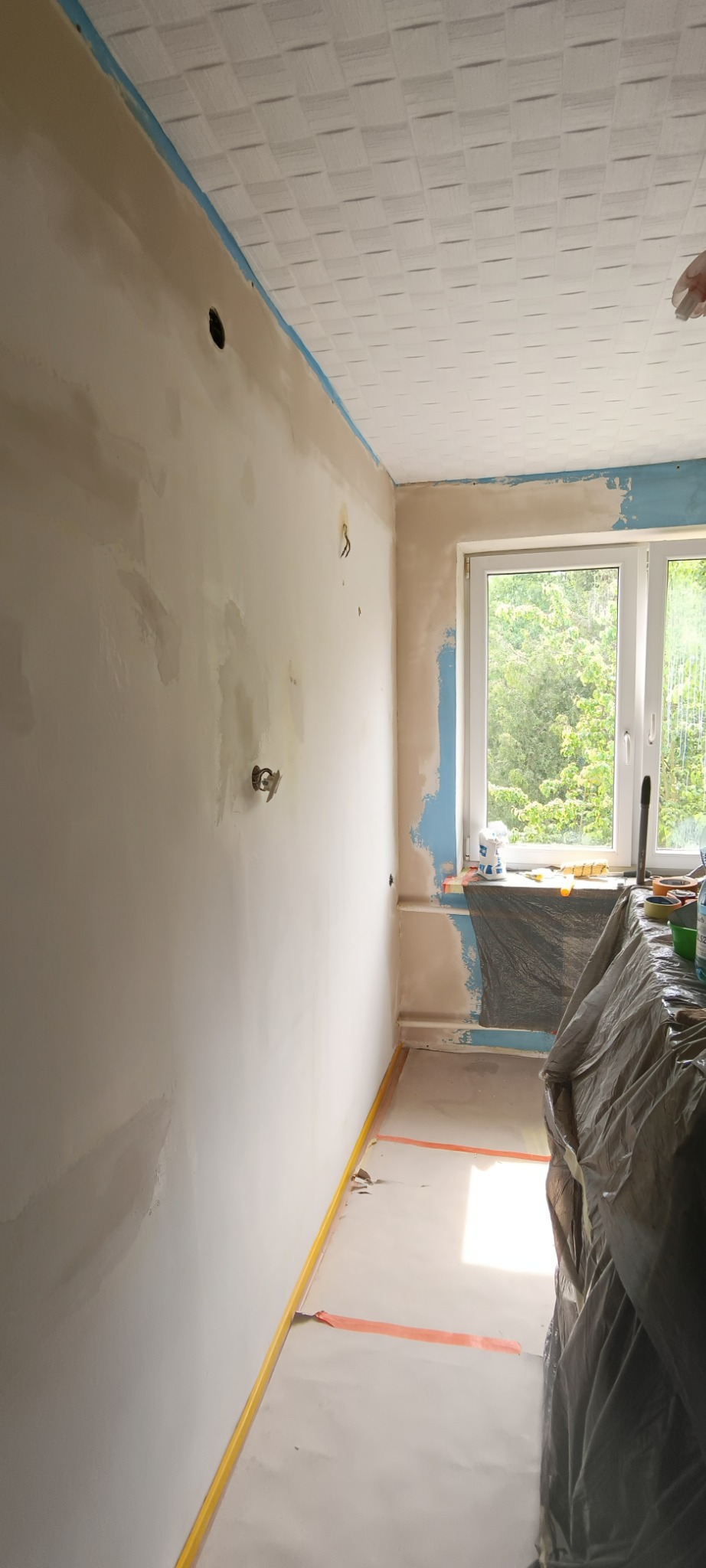 Pokój w trakcie malowania: ściany częściowo pokryte gładzią, zabezpieczone okno i podłoga folią malarską, widoczne puszki z farbą i narzędzia.