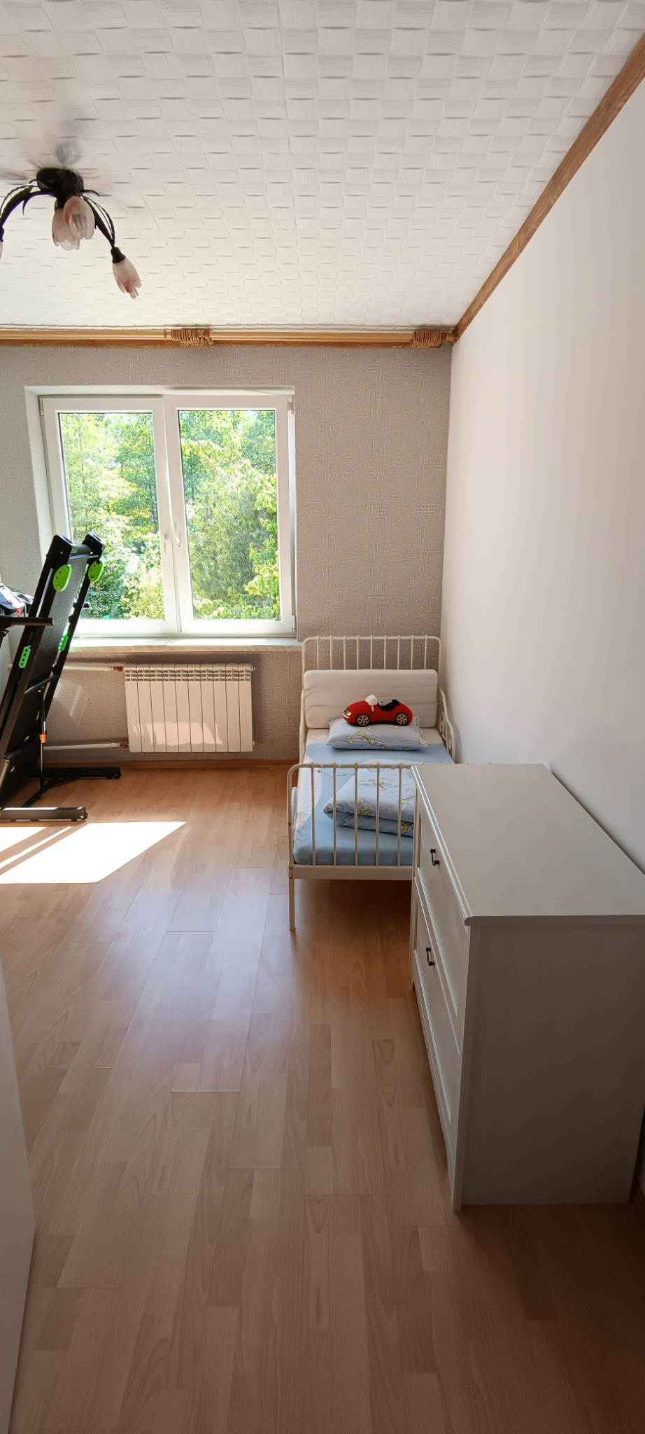 Jasny pokój dziecięcy z drewnianą podłogą, białymi meblami i tapetą w neutralnym odcieniu, widok na zewnątrz przez okno.