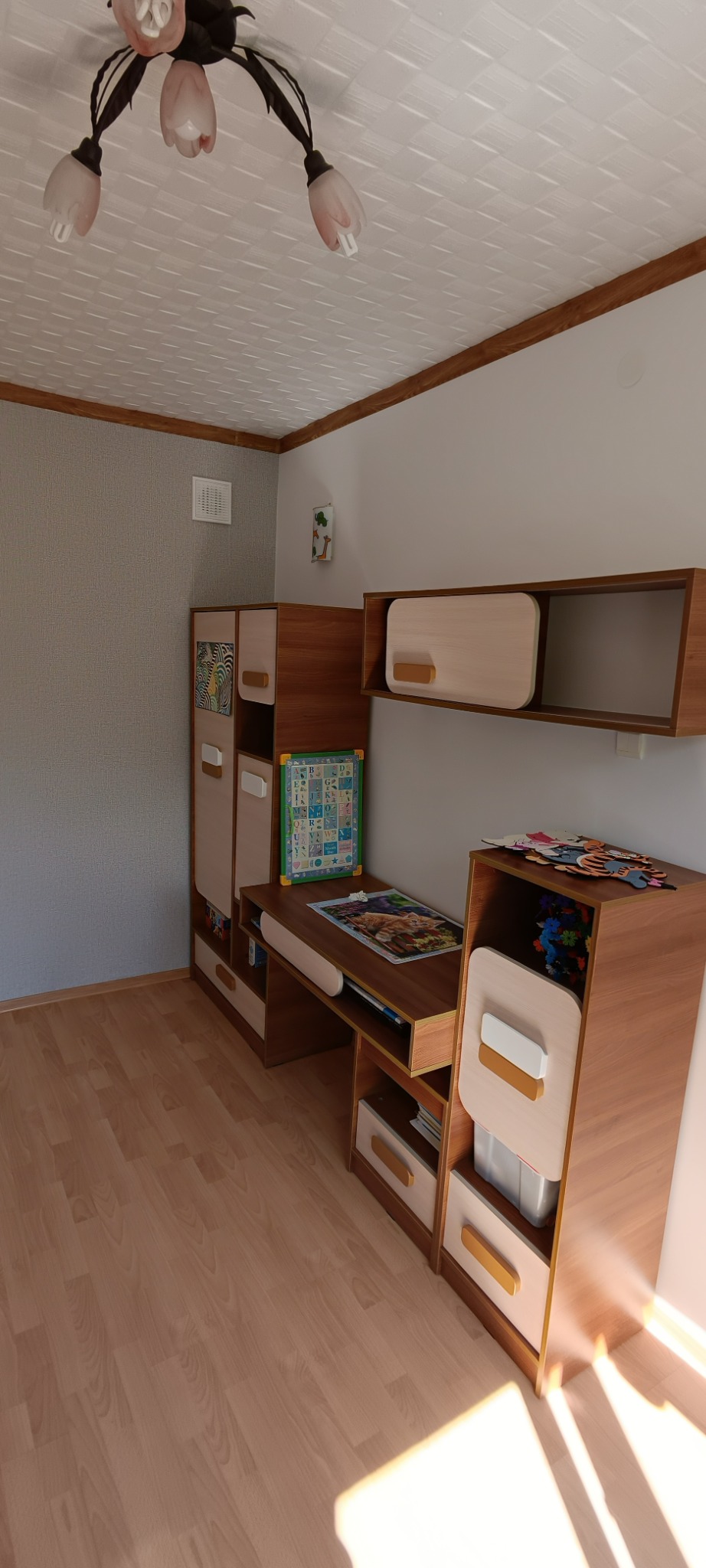 Pokój dziecięcy z drewnianymi panelami podłogowymi, meblami z jasnego drewna i białymi akcentami, w tym biurkiem z półkami, szafkami i lampą sufitową z różowymi kloszami.