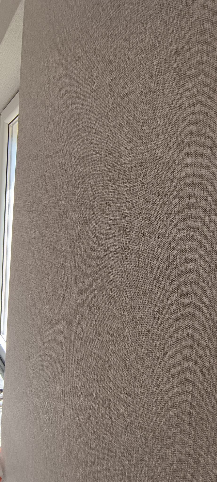 Beżowa ściana pokryta teksturowaną tapetą w pobliżu okna z widocznym fragmentem ramy.