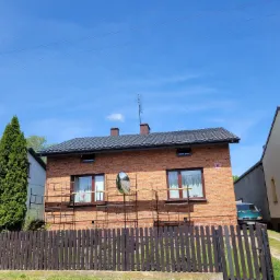 Dom z ciemną dachówką i ceglaną elewacją, na tle błękitnego nieba, z widocznymi rusztowaniami i lustrem drogowym przed budynkiem.