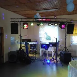 Rozstawiony sprzęt nagłośnieniowy i oświetleniowy na sali z dekoracjami balonowymi, widoczne kolumny głośnikowe, keyboard, statywy mikrofonowe, oświetlenie sceniczne z napisem 'PRODUKC'