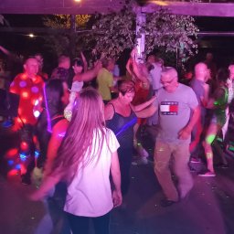 Nikoletta Żmuda - Roztańczony tłum pod drewnianą konstrukcją oświetloną kolorowymi światłami, widoczne drzewa w tle.