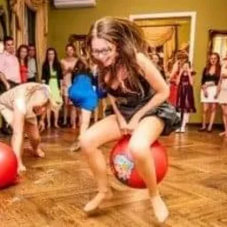 Dziewczyna w okularach skacze na czerwonej piłce do skakania z nadrukiem w pomieszczeniu z parkietem i widzami w tle, podczas zabawy z animatorem.