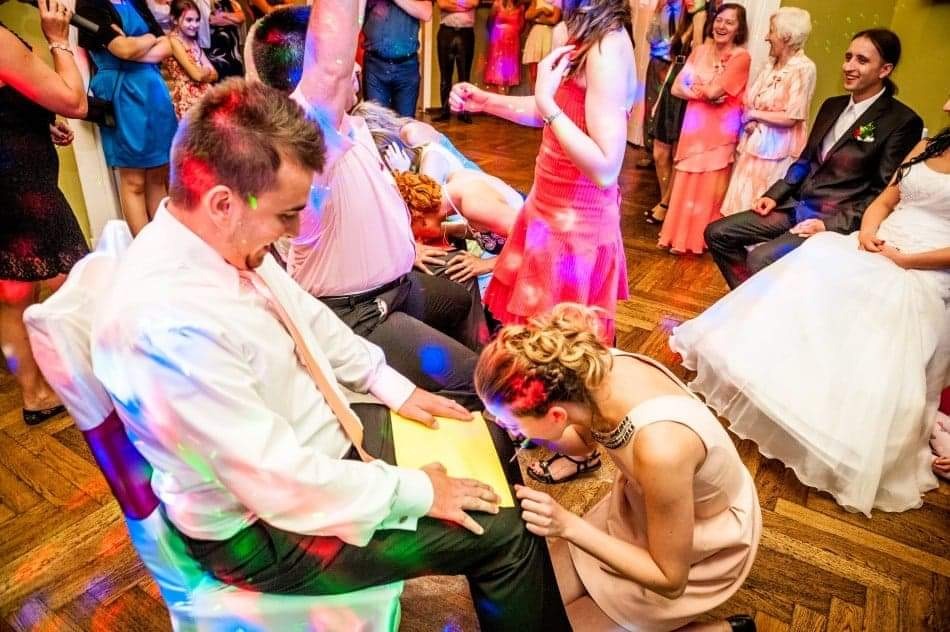 Panna młoda klęczy przed panem młodym podczas oczepin, w tle goście weselni, parkiet i efekty świetlne.