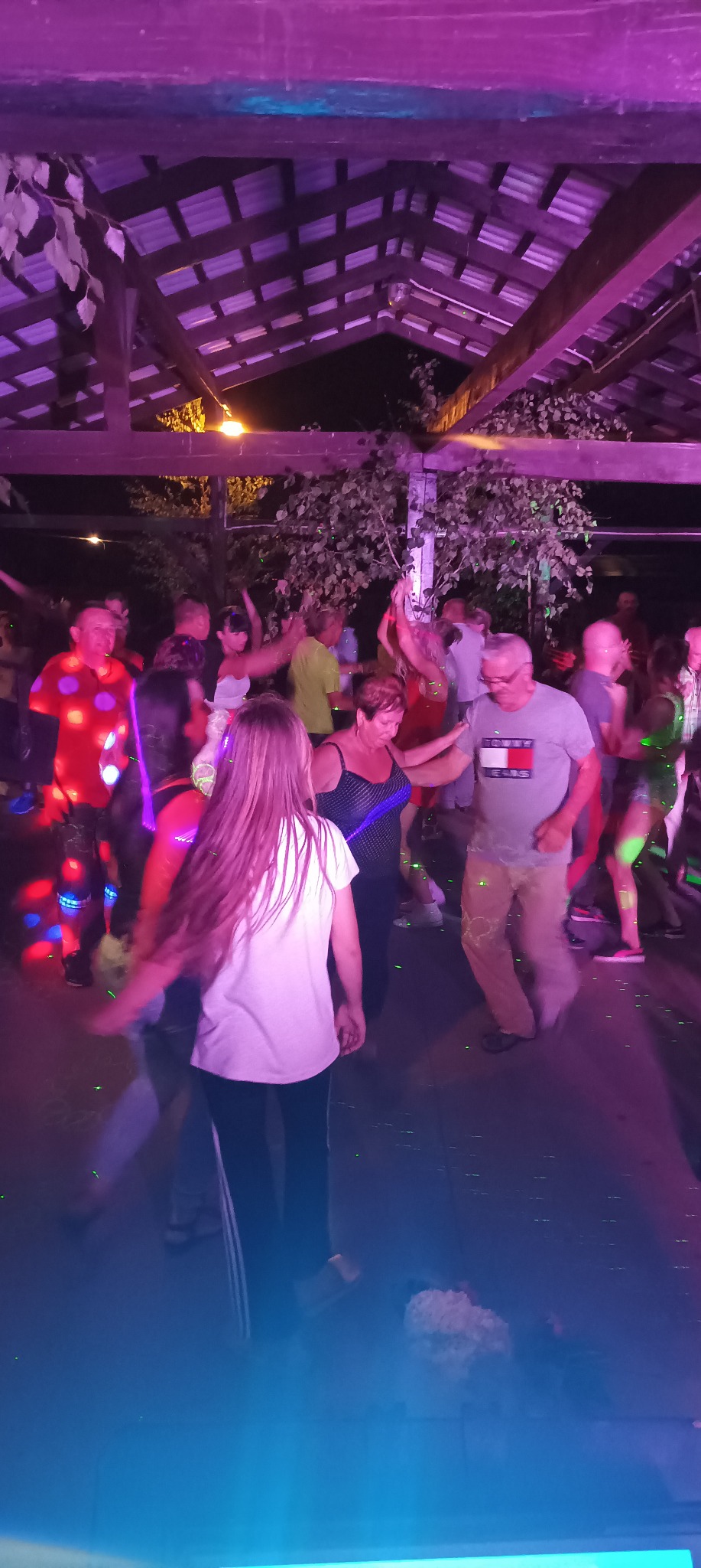 Roztańczony tłum pod drewnianą konstrukcją oświetloną kolorowymi światłami, widoczne drzewa w tle.