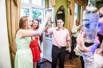 Mężczyzna w różowej koszuli wznosi toast kieliszkiem na tle rozbawionych gości podczas uroczystości.