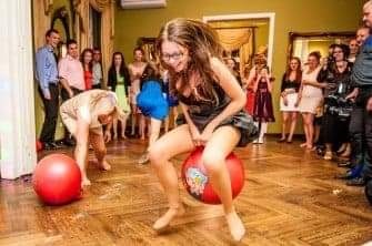 Dziewczyna w okularach skacze na czerwonej piłce do skakania z nadrukiem w pomieszczeniu z parkietem i widzami w tle, podczas zabawy z animatorem.