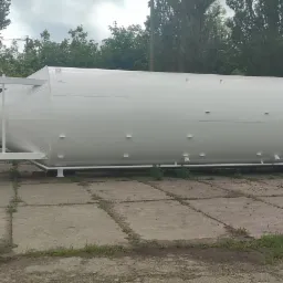 Duży, poziomy, biały silos przemysłowy z metalową konstrukcją wsporczą i panelem sterowania, stojący na betonowym placu z zielenią w tle.