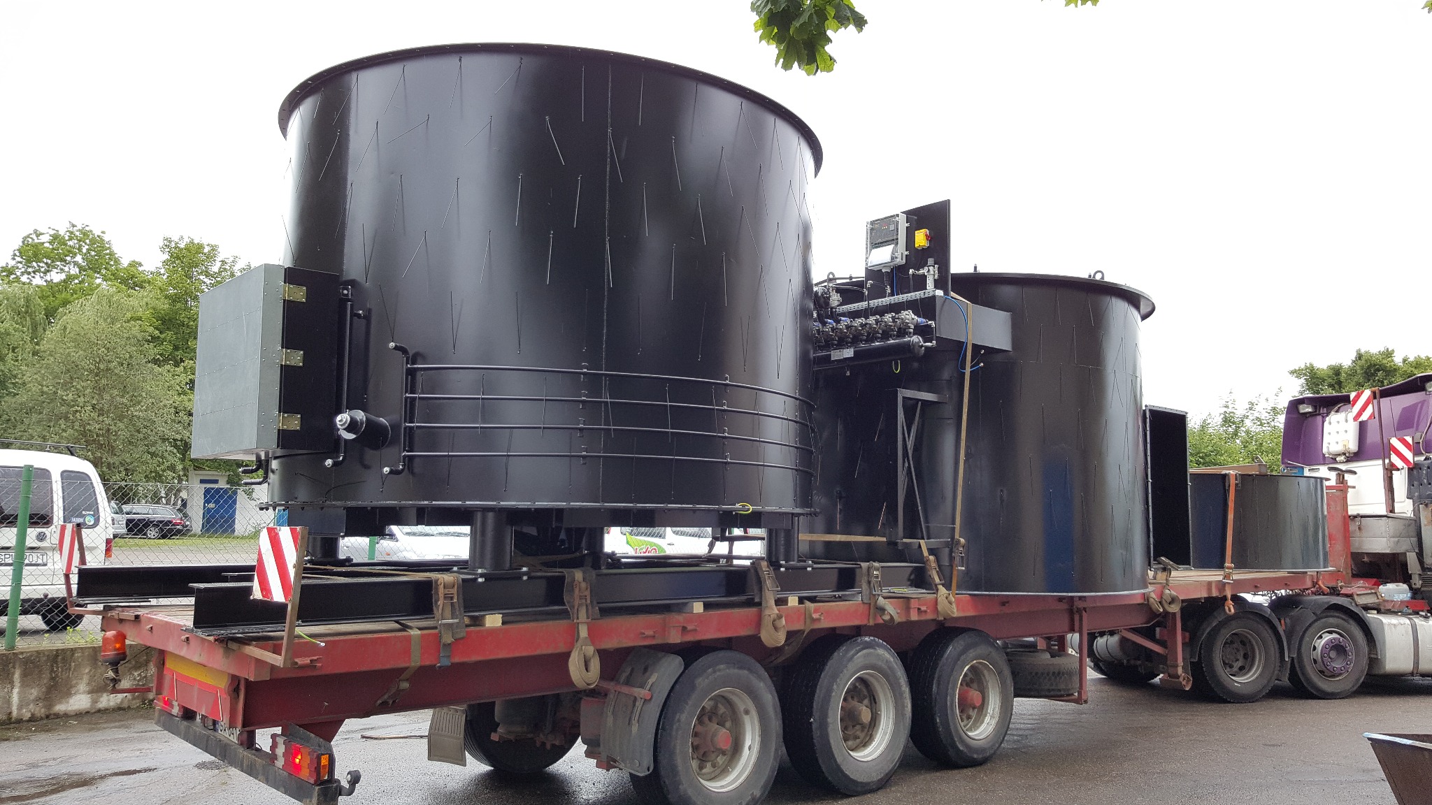 Dwa duże, czarne zbiorniki przemysłowe z metalu, przewożone na platformie ciężarówki z czerwonymi elementami zabezpieczającymi, widoczne na zewnątrz budynku z zielenią w tle.