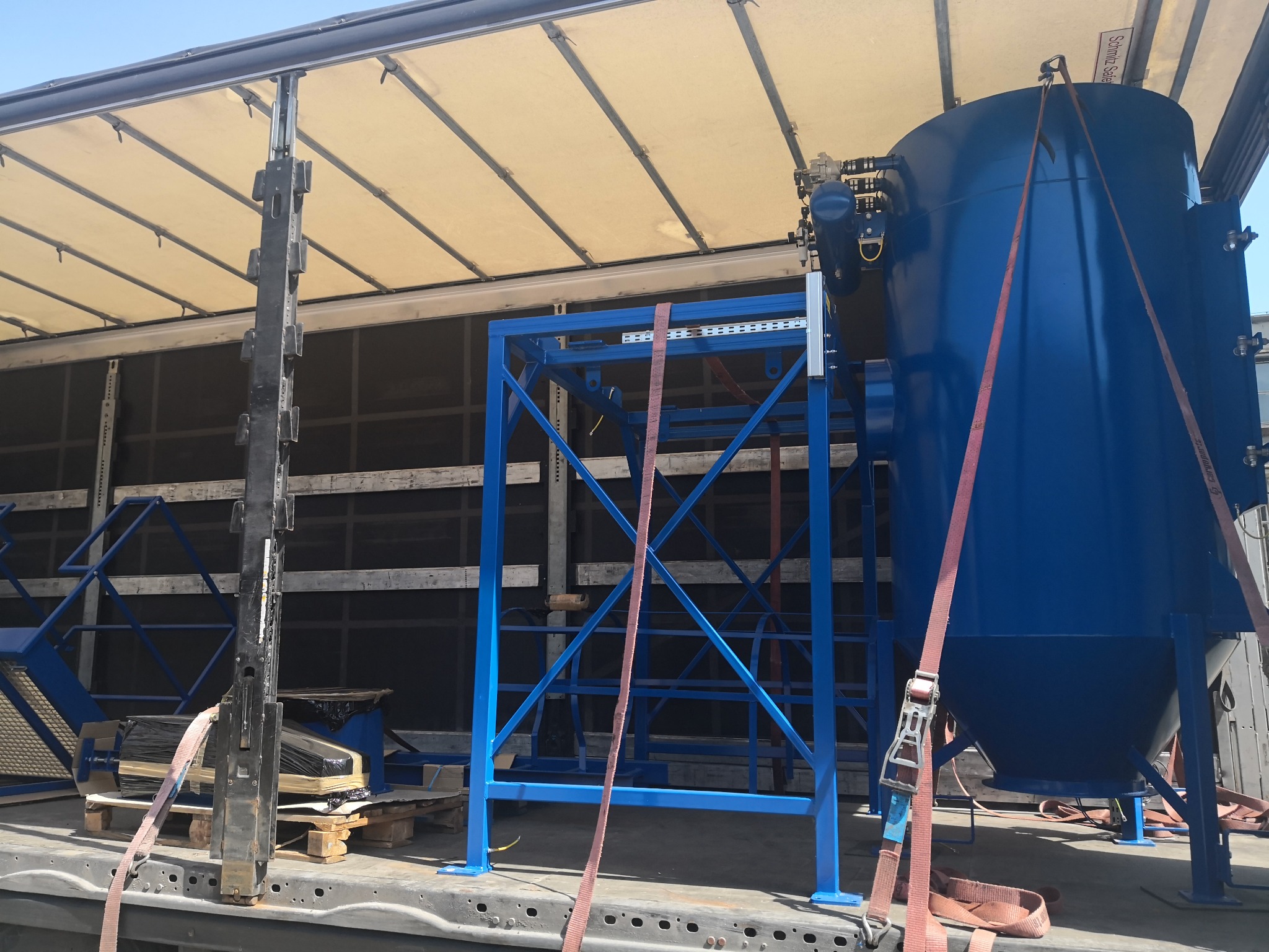 Niebieski zbiornik przemysłowy i stalowa konstrukcja zabezpieczone pasami transportowymi wewnątrz naczepy ciężarówki, widoczne elementy mocujące i podkłady drewniane.