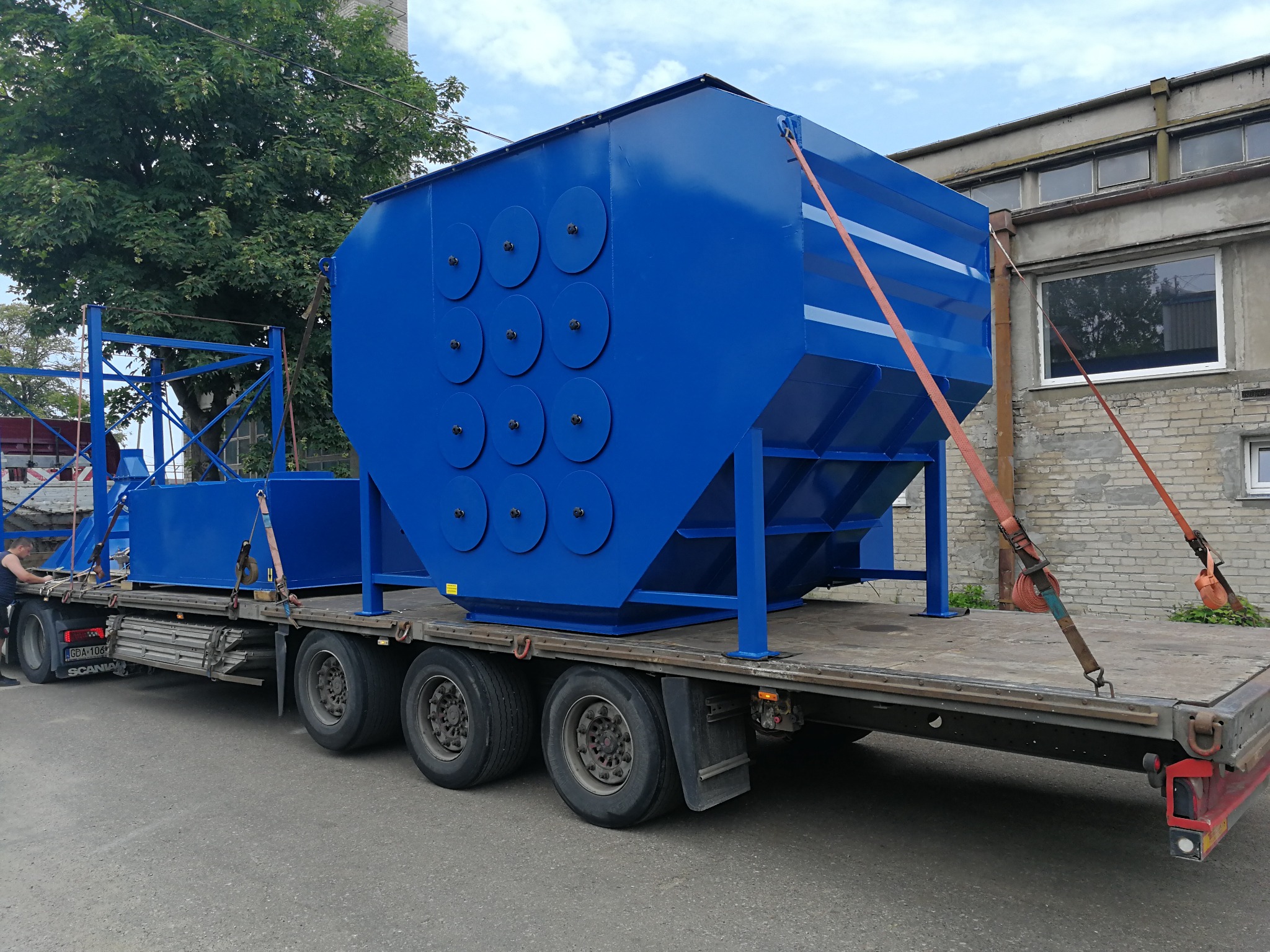 Niebieski zbiornik przemysłowy z okrągłymi wzmocnieniami bocznymi, transportowany na naczepie ciężarówki, zabezpieczony pomarańczowymi pasami.