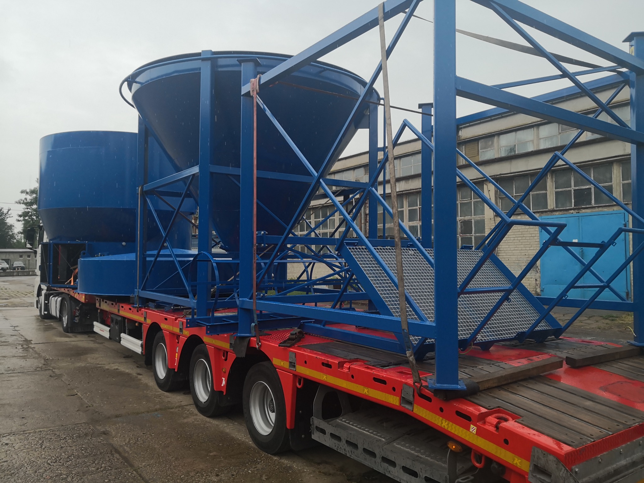 Transport dwóch niebieskich silosów na czerwonej platformie ciężarówki w Pucku, widoczne stalowe konstrukcje zabezpieczające ładunek i fragment budynku w tle.