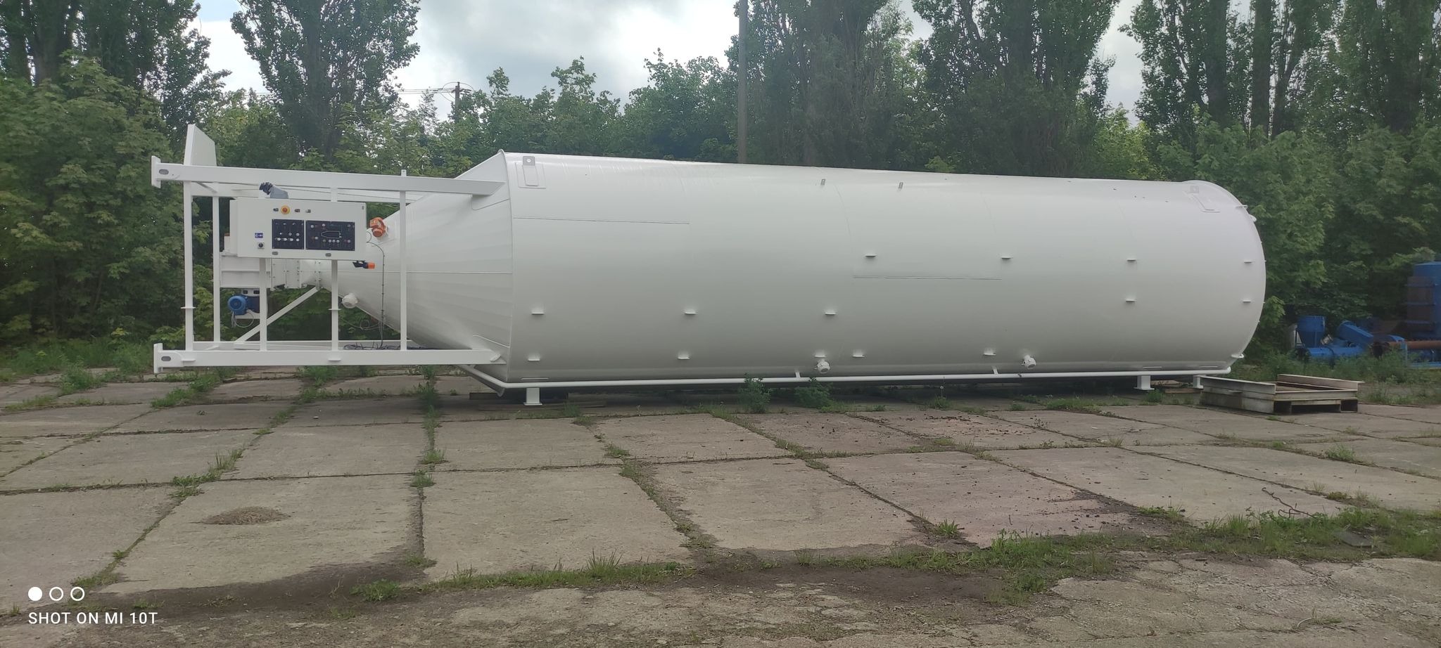 Duży, poziomy, biały silos przemysłowy z metalową konstrukcją wsporczą i panelem sterowania, stojący na betonowym placu z zielenią w tle.