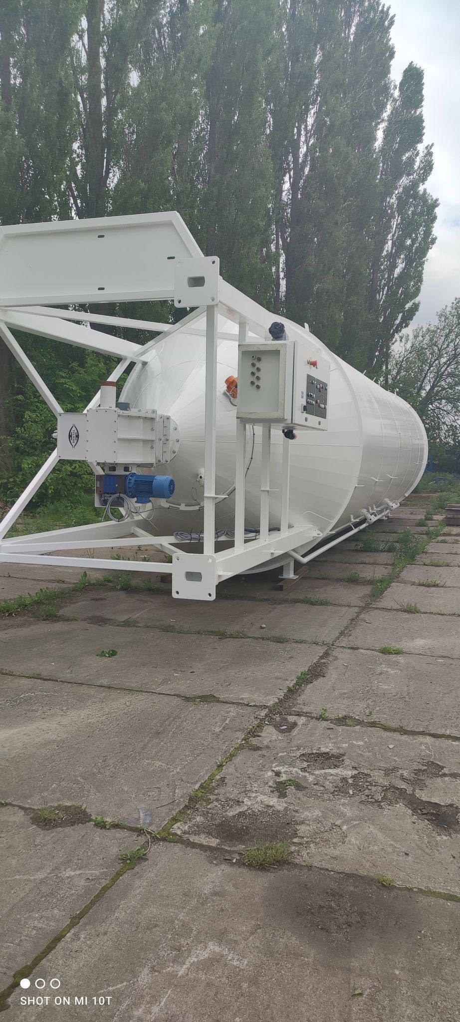 Biały, cylindryczny silos na stalowej konstrukcji, umieszczony na betonowej nawierzchni, z widocznymi elementami sterującymi i silnikiem.