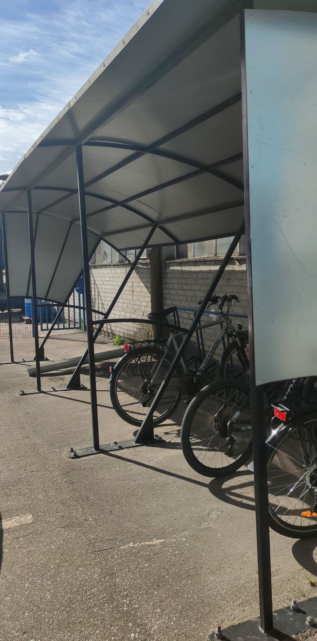 Zadaszony parking rowerowy z metalową konstrukcją i szarym dachem, rowery zaparkowane wewnątrz, ceglany budynek w tle.