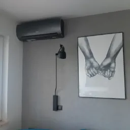 Czarny klimatyzator ścienny zamontowany w nowoczesnym salonie z szarą ścianą, minimalistyczną lampą i obrazem dłoni.